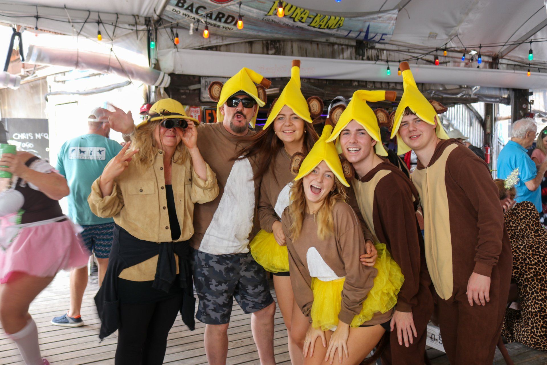 A group of people wearing banana costumes and hats poses for a photo under a decorated outdoor tent canopy.