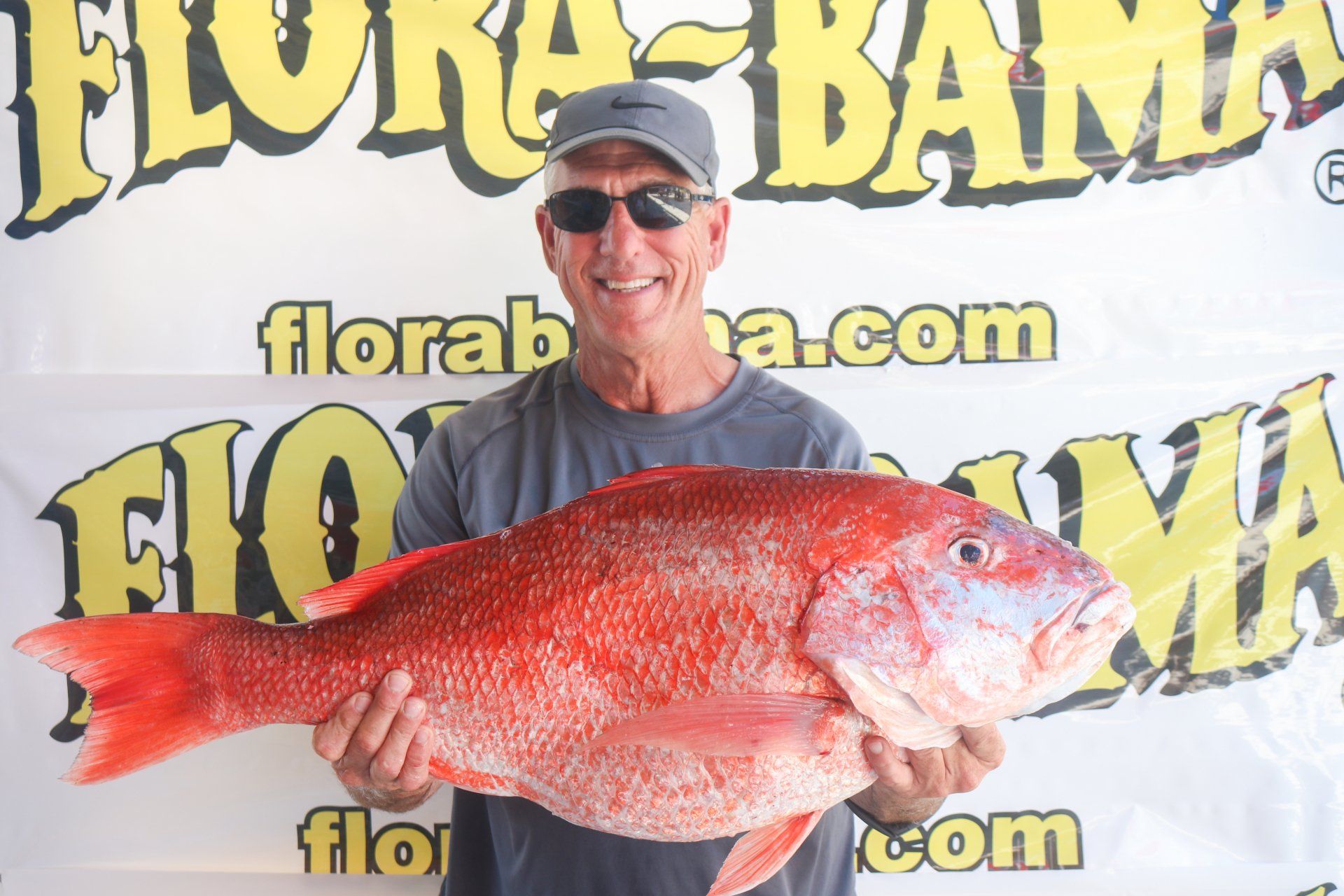 A person wearing sunglasses and a cap holds a large red snapper fish in front of a Flora-Bama branded backdrop.