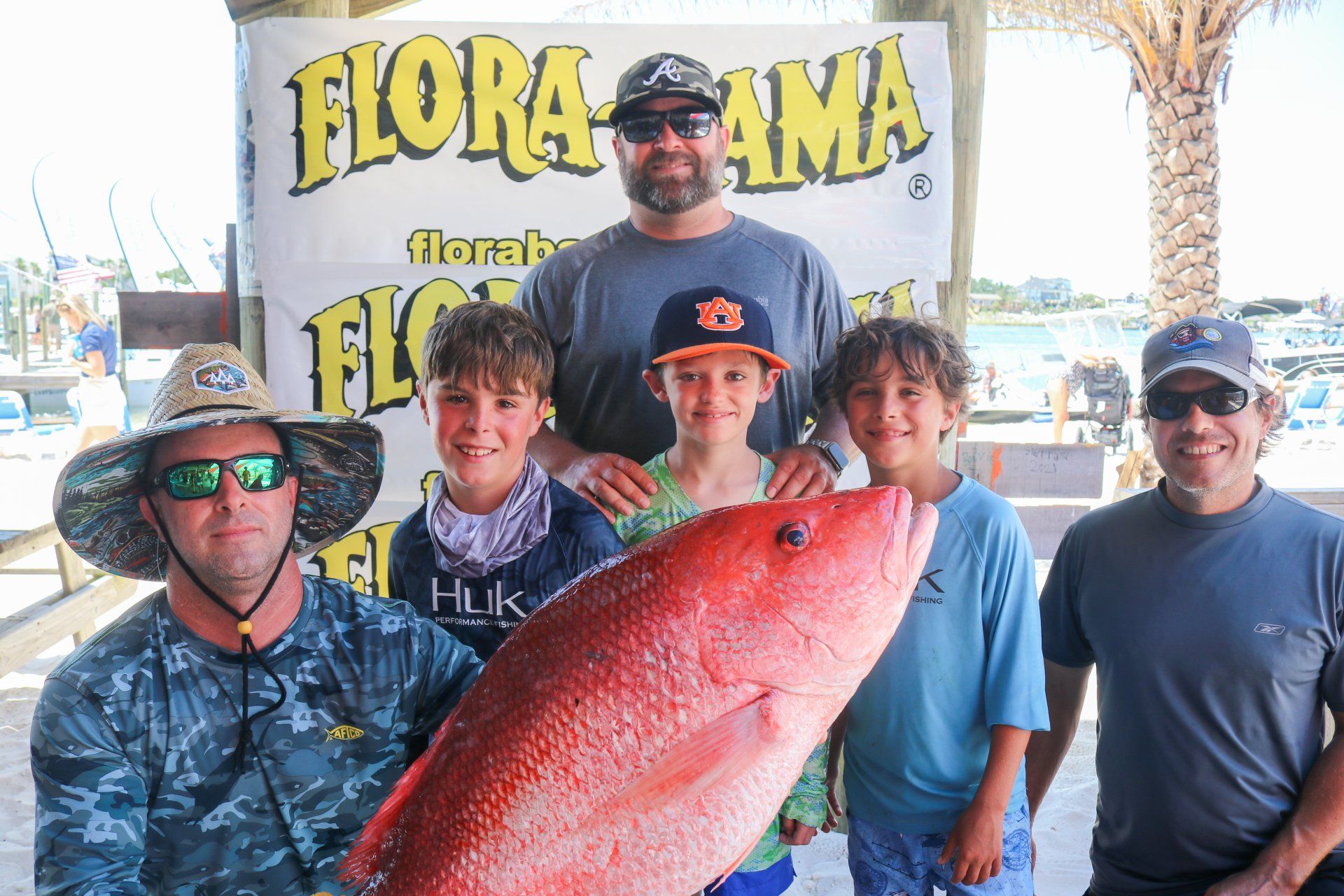 Group of six people standing outdoors in front of a Flora-Bama banner, holding a large red fish for a fishing event.