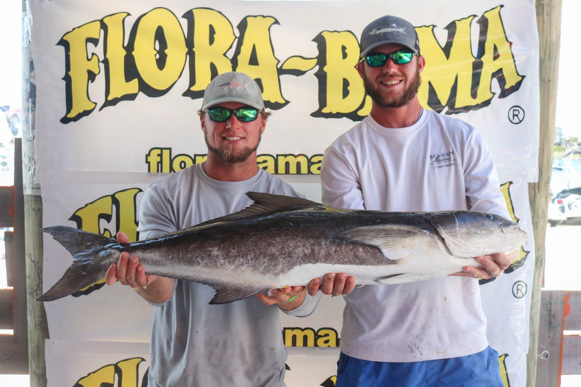 Two people stand in front of a Flora-Bama sign holding a large, dark fish caught during a fishing competition.