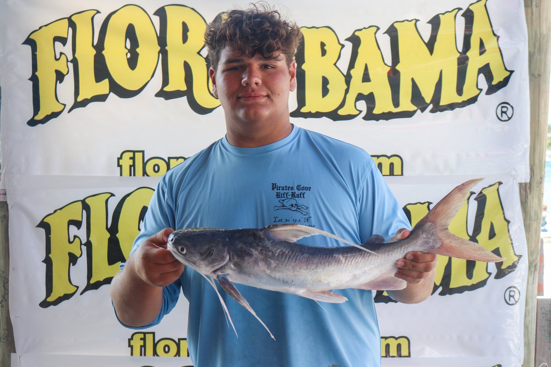 A young person holds a silver saltwater catfish in front of a Flor-Bama branded backdrop.