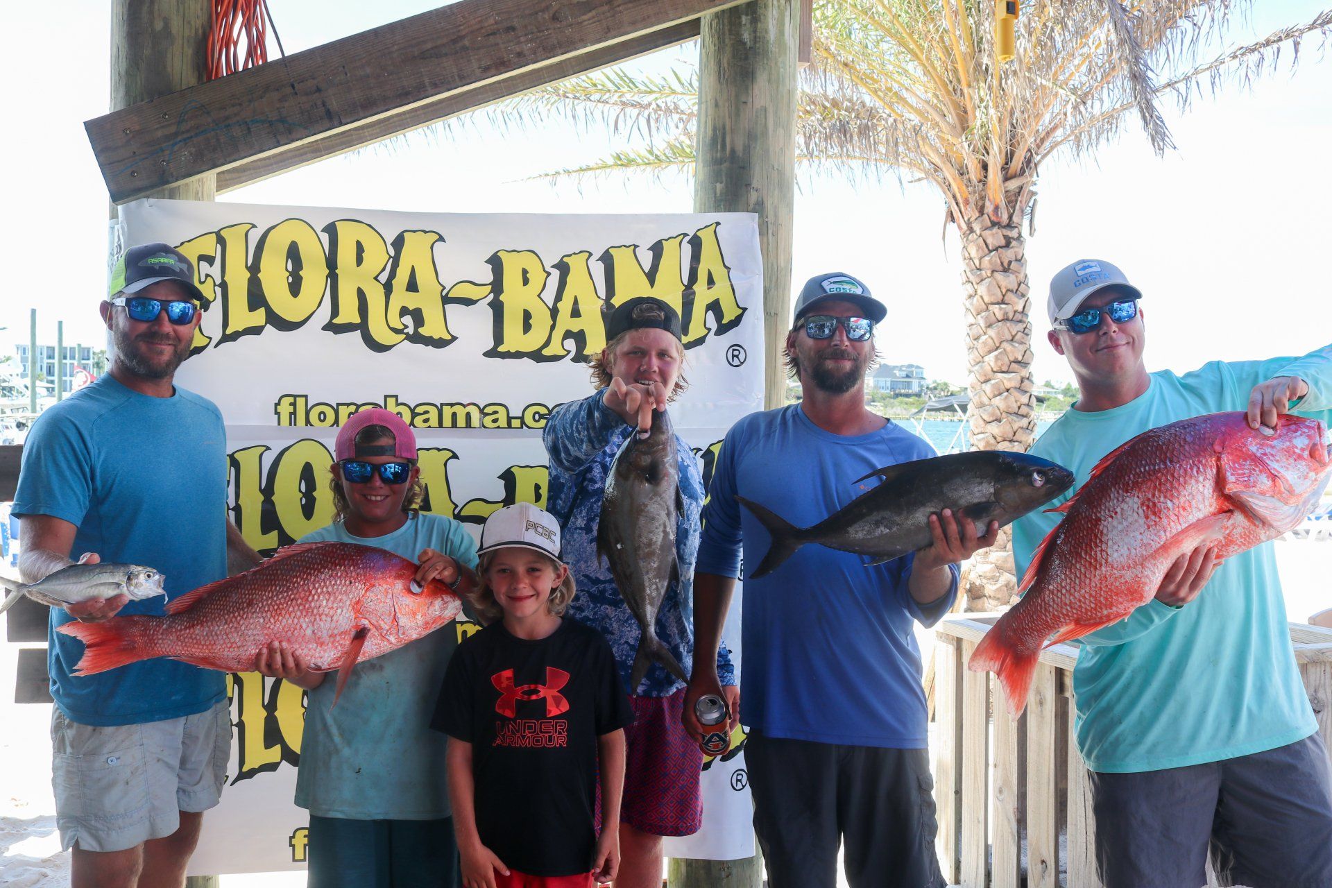 Five people holding caught fish in front of a Flora-Bama banner on a sunny day.