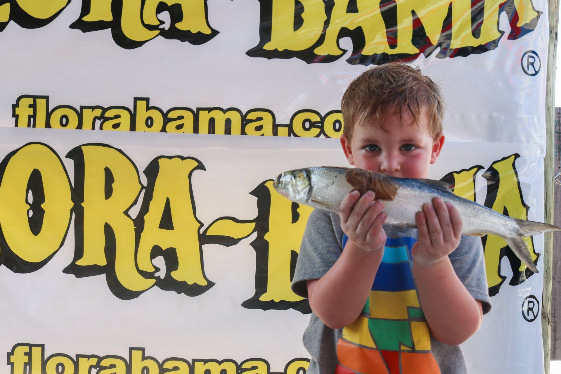 A child holds a fish in front of their face, standing in front of a yellow and white Florabama banner.