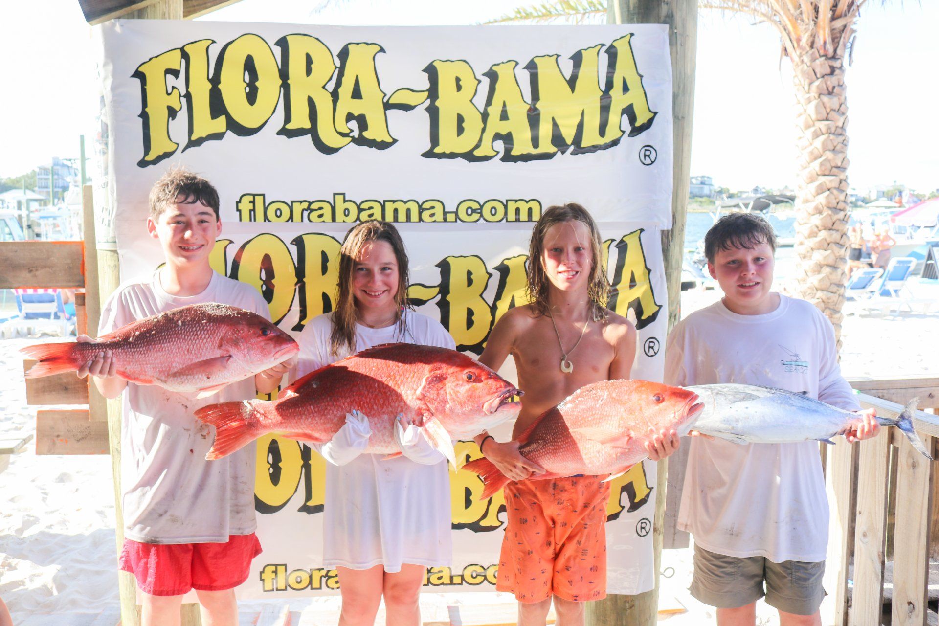 Four people stand in front of a Flora-Bama sign at a beach, each holding a large, freshly caught red snapper.