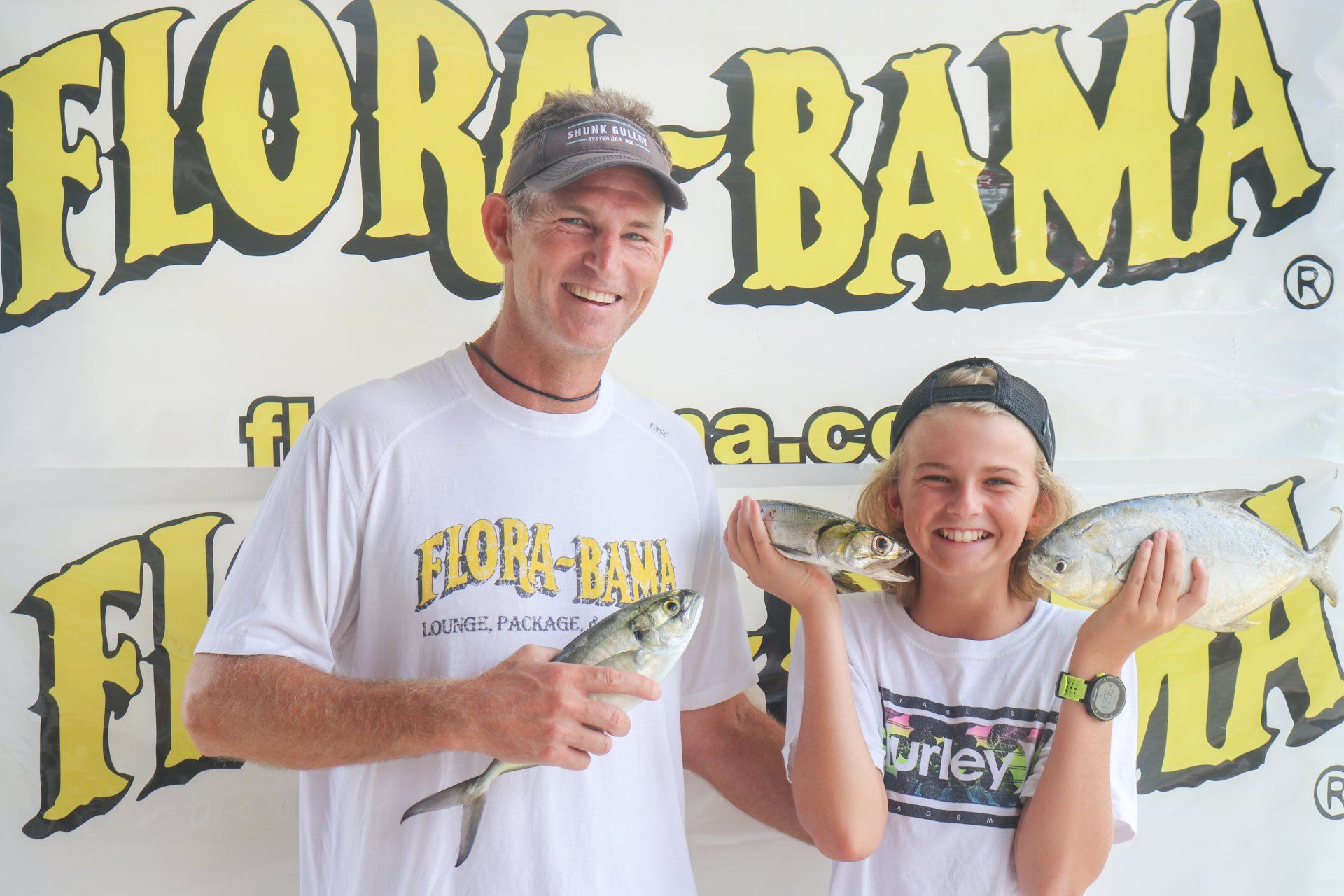 A smiling adult and youth hold fish in front of a Flora-Bama branded backdrop.