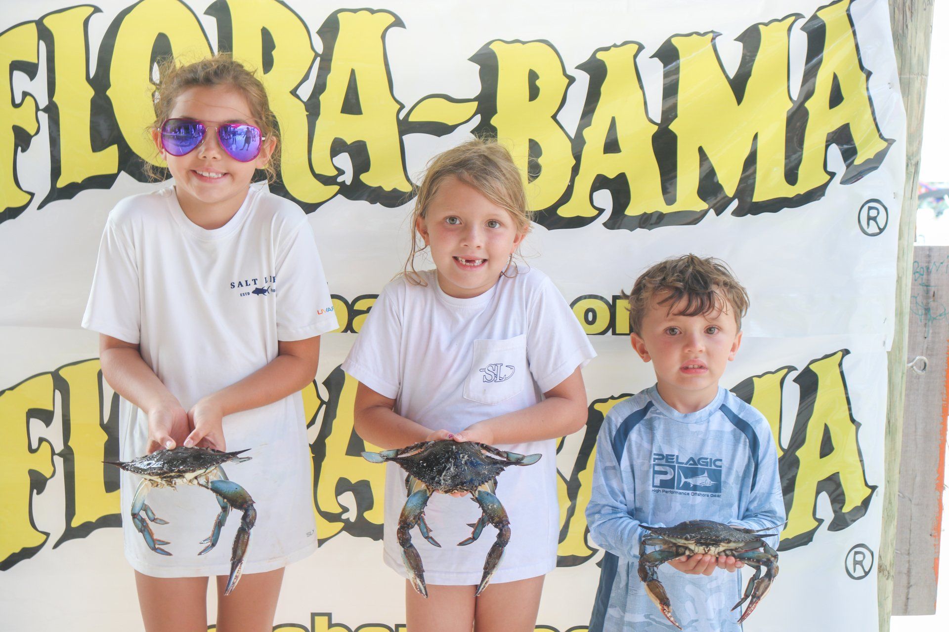 Three children stand before a Flora-Bama sign, each holding a blue crab.