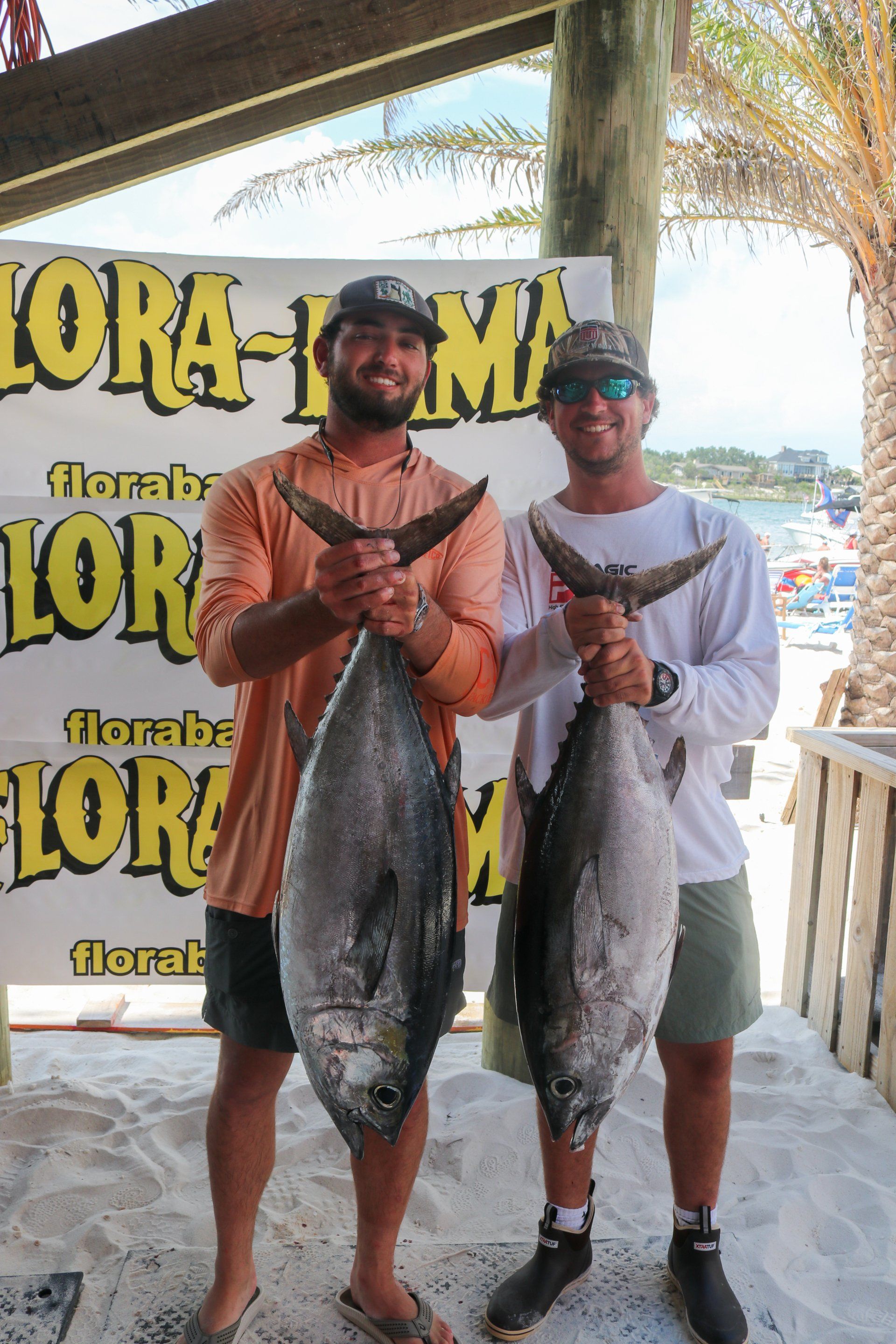 Two people holding large tuna fish in front of a Florabama beach sign, standing on sand under a shaded structure.