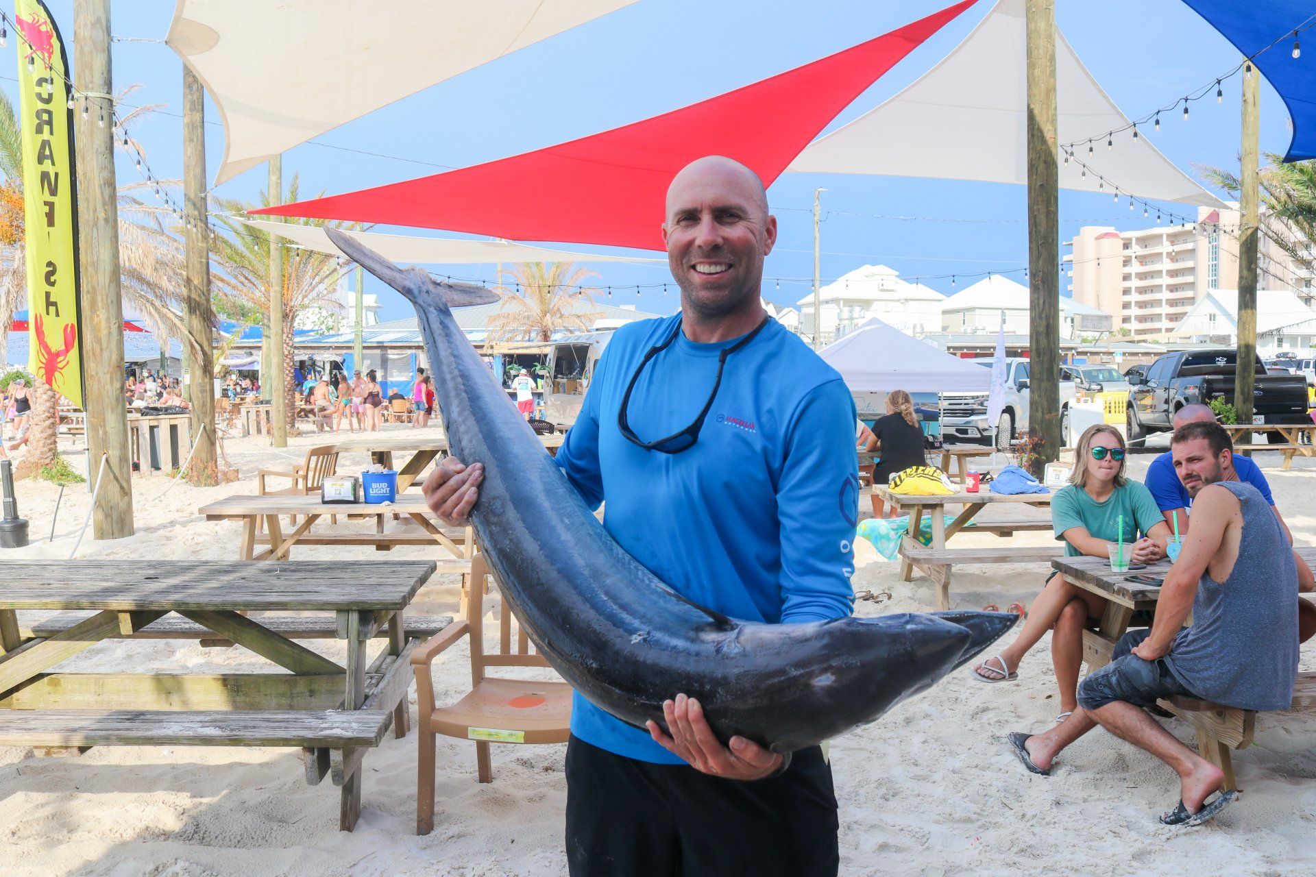 A person smiles while holding a large fish on a sandy beach under colorful shade sails, with tables and people nearby.