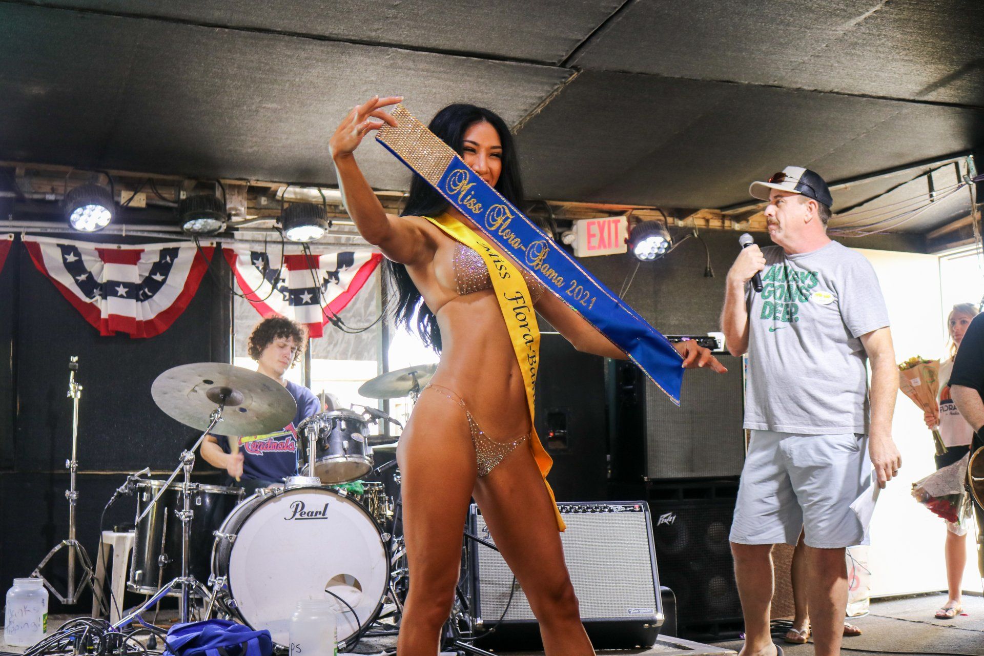 A woman wearing a swimsuit and beauty pageant sash poses on a stage, while a drummer performs and a man holds a microphone.