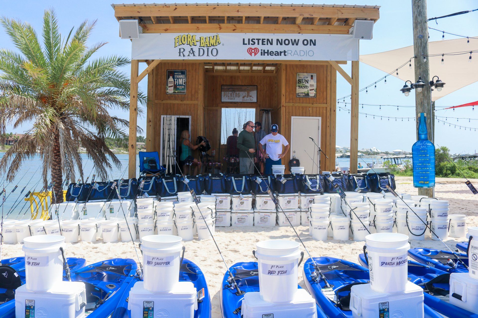 Rows of white buckets and blue kayaks lined up on a beach in front of a wooden radio station booth under a clear sky.