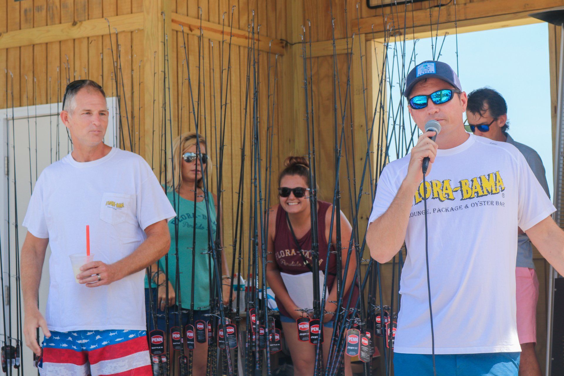A person speaks into a microphone while others stand in front of numerous fishing rods inside a wooden structure.