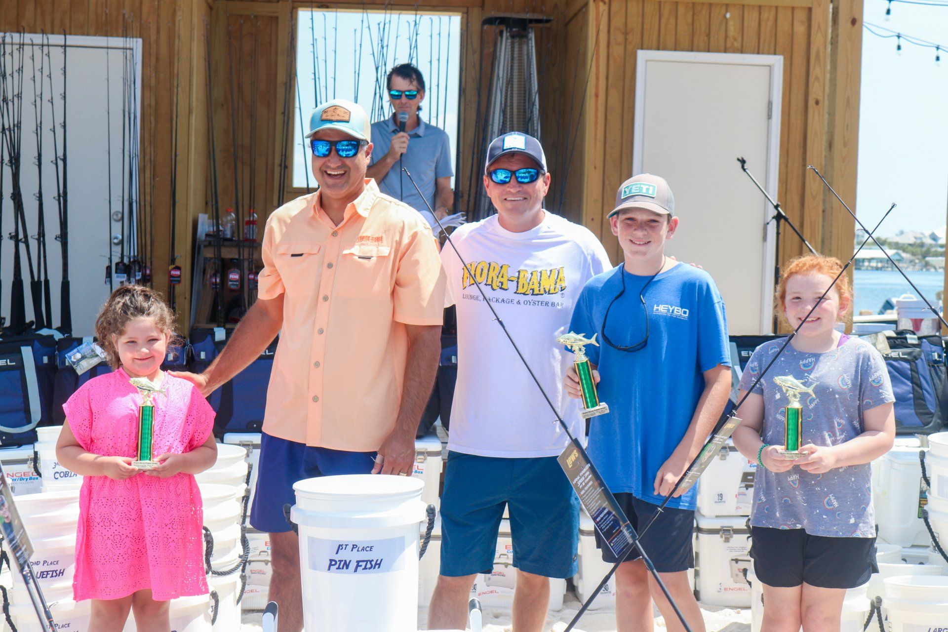 Four people stand on a dock holding small trophies, with a man in the background, in an outdoor fishing tournament setting.