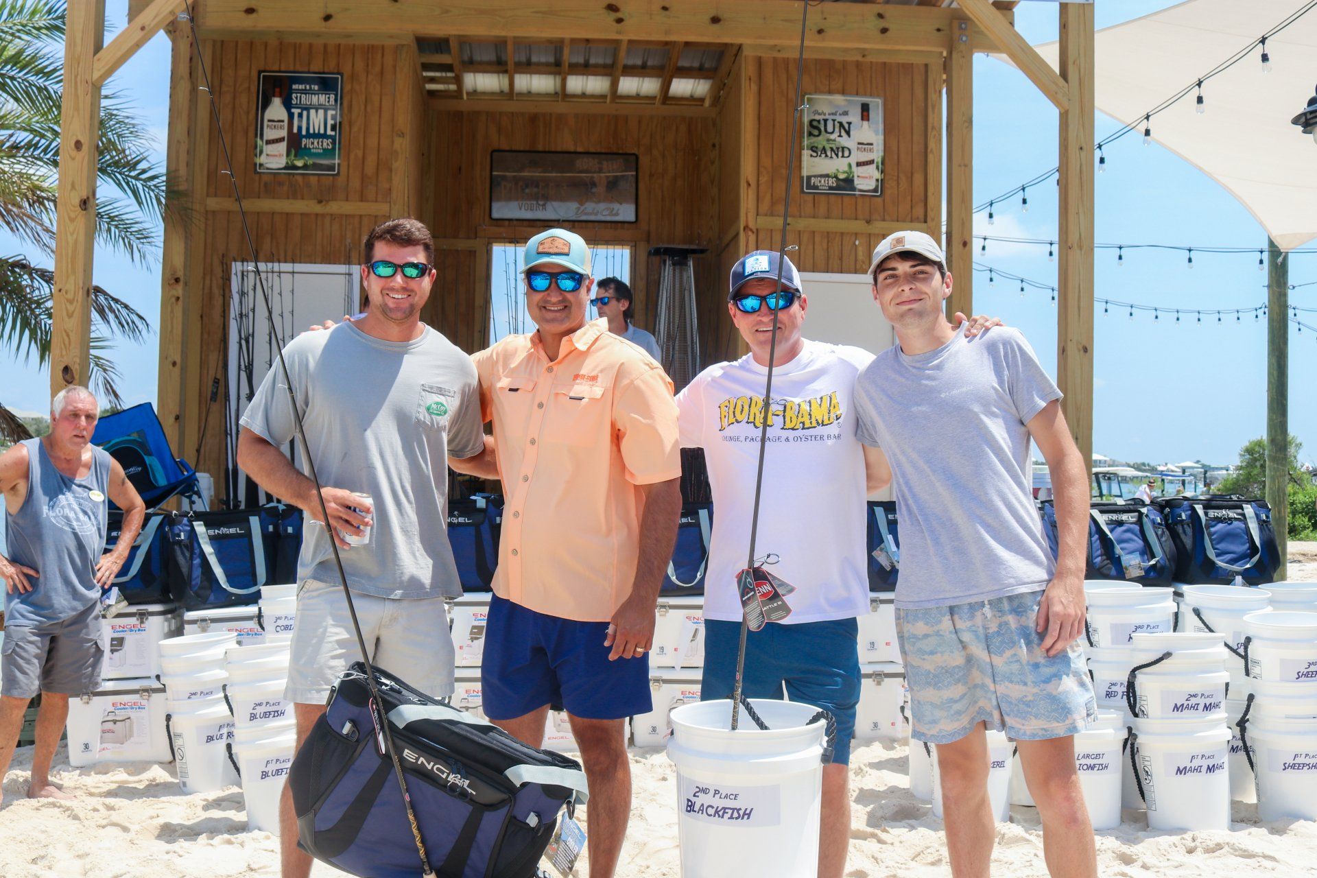 Four people in casual beach attire pose for a photo in front of a wooden beach structure with many white buckets nearby.