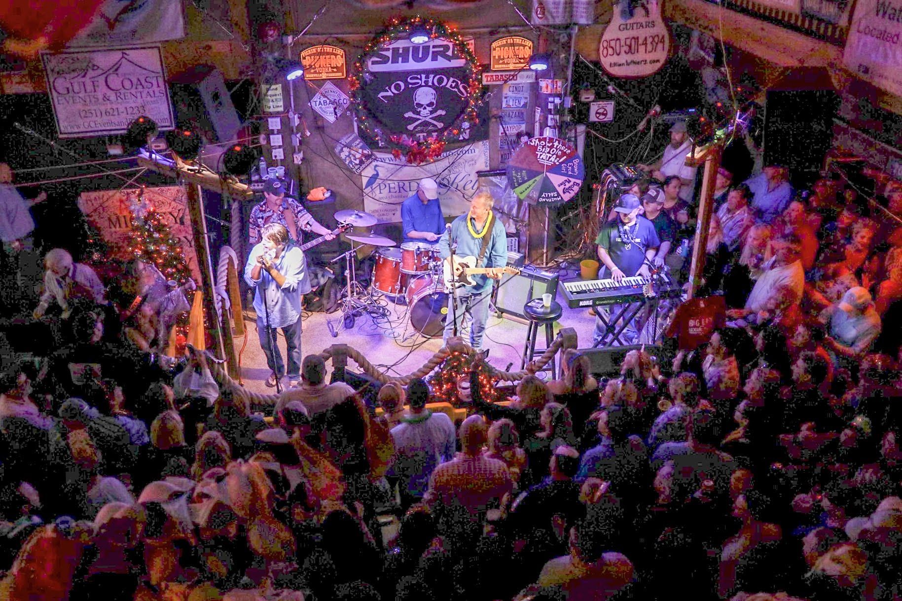 A band performs on a stage decorated with skull imagery in a crowded, dimly lit venue with colorful stage lights.