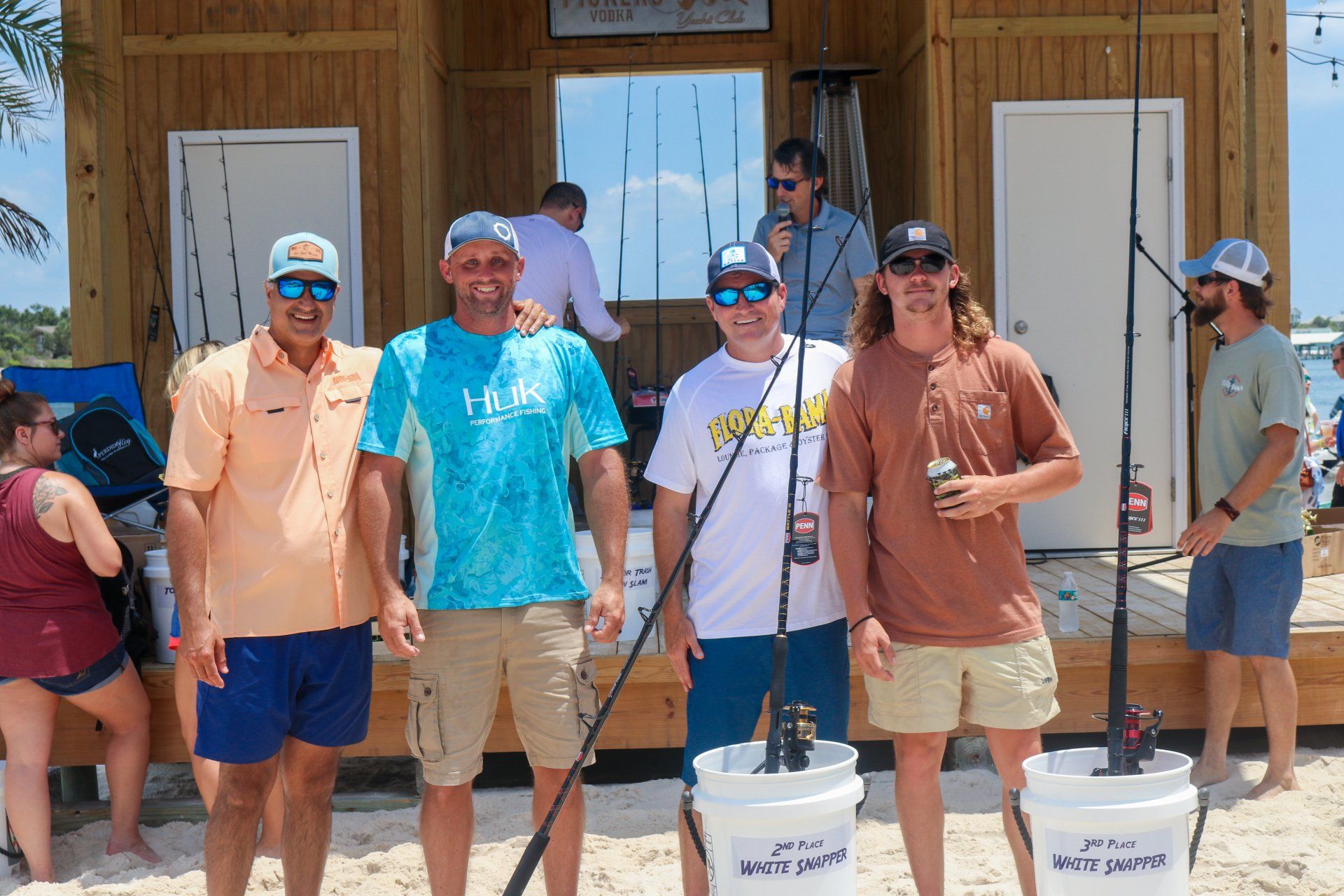 Four people stand in a line on a beach in front of a wooden structure, with two fishing rods held in white buckets.