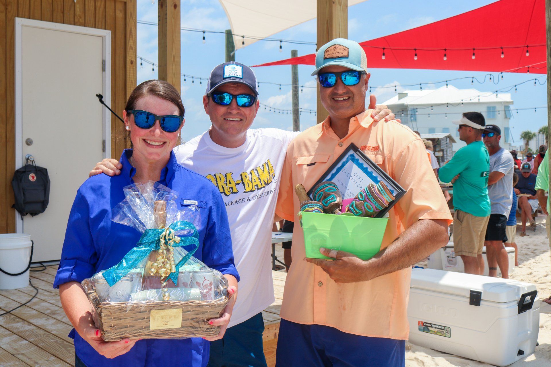 Three smiling people holding gift baskets and awards outdoors under a red sunshade at a beach event.
