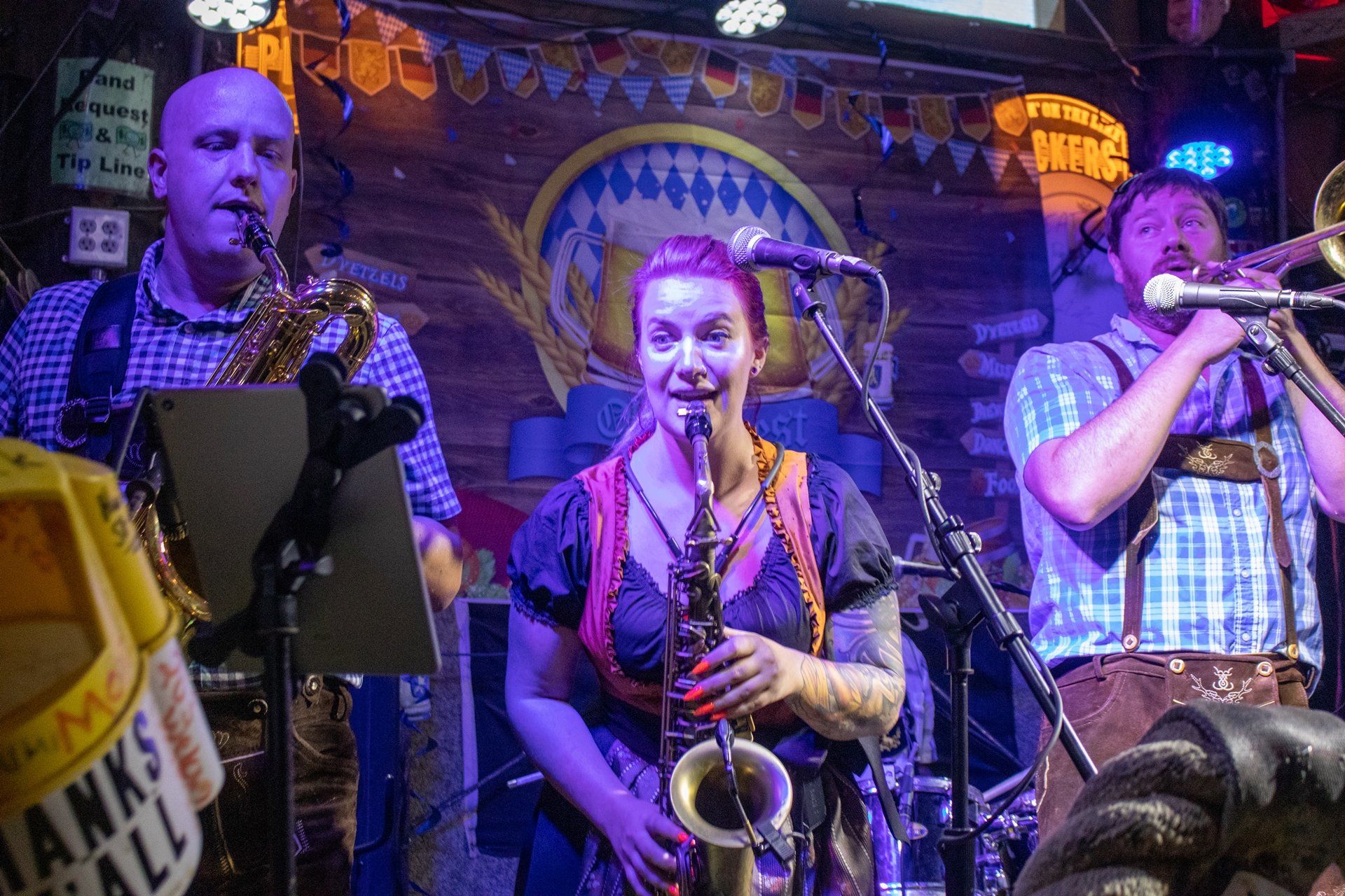 A band performs on stage in Bavarian-style clothing, featuring two saxophone players and a trombone player.