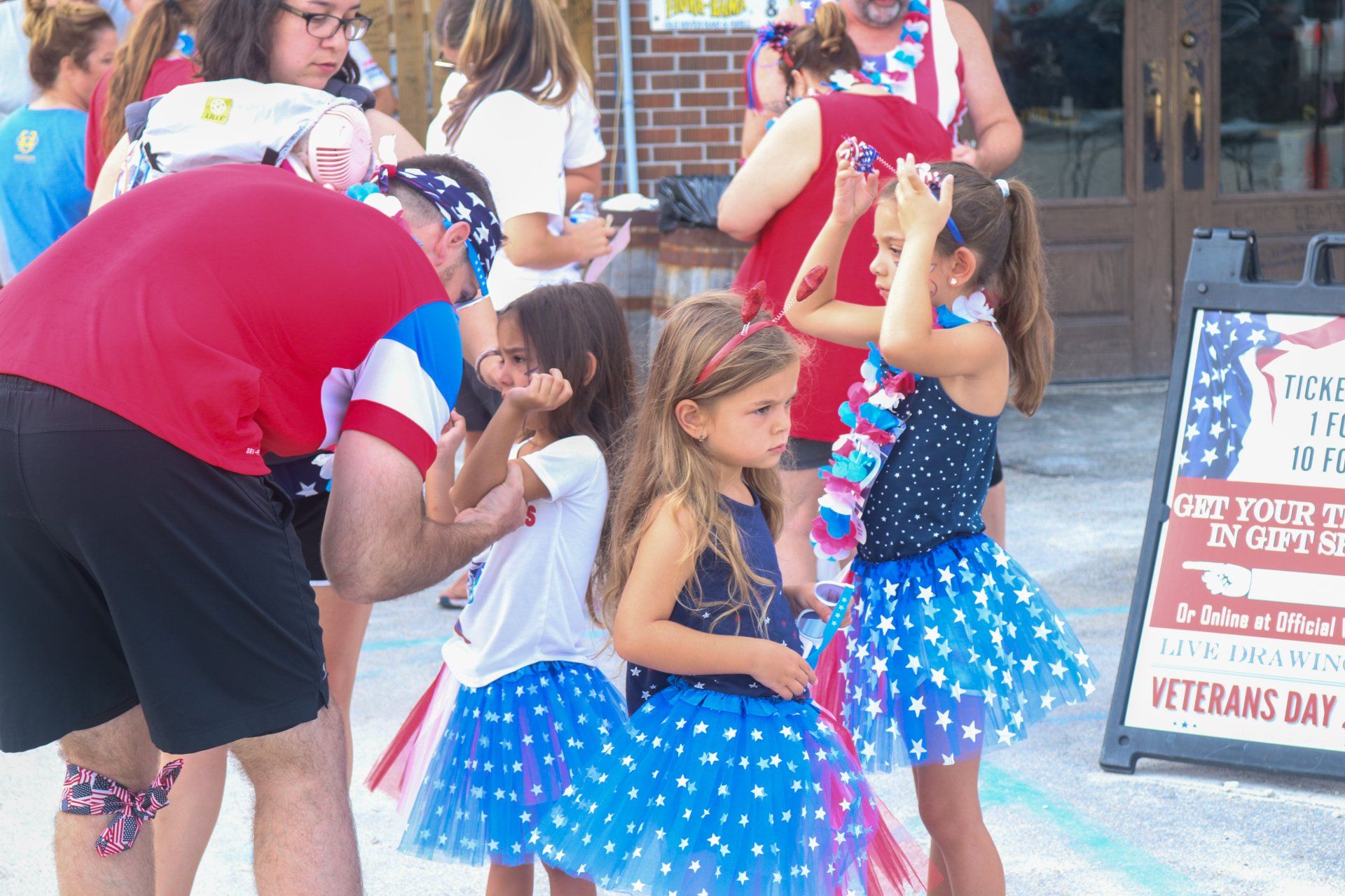 Children wearing star-patterned blue skirts and red, white, and blue decorations stand near an event sign on a sidewalk.