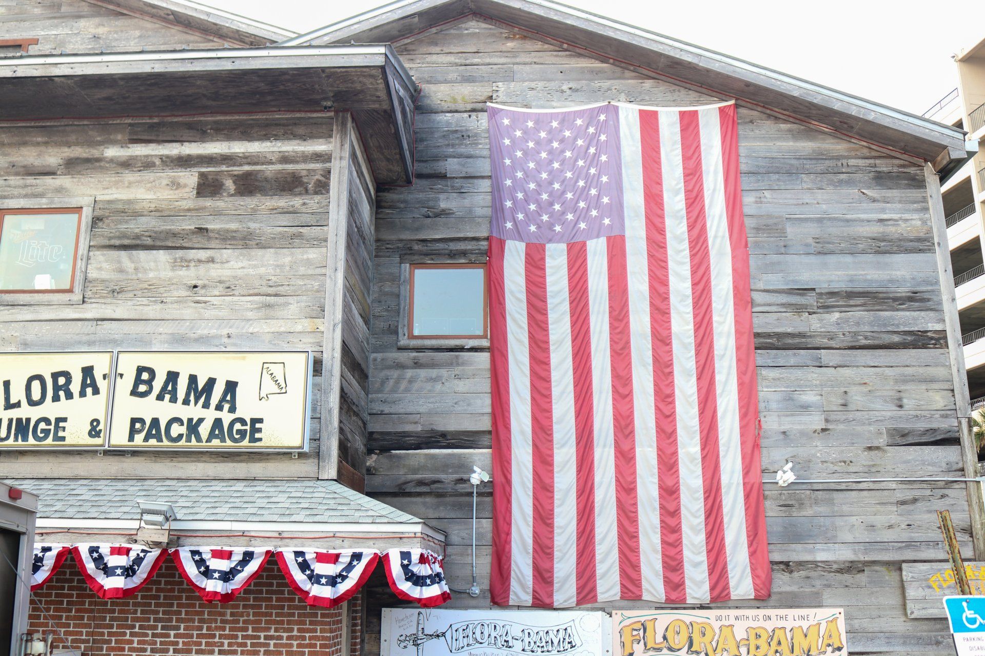 A large American flag hangs on the wooden exterior of the Flora-Bama lounge, adorned with red, white, and blue bunting.