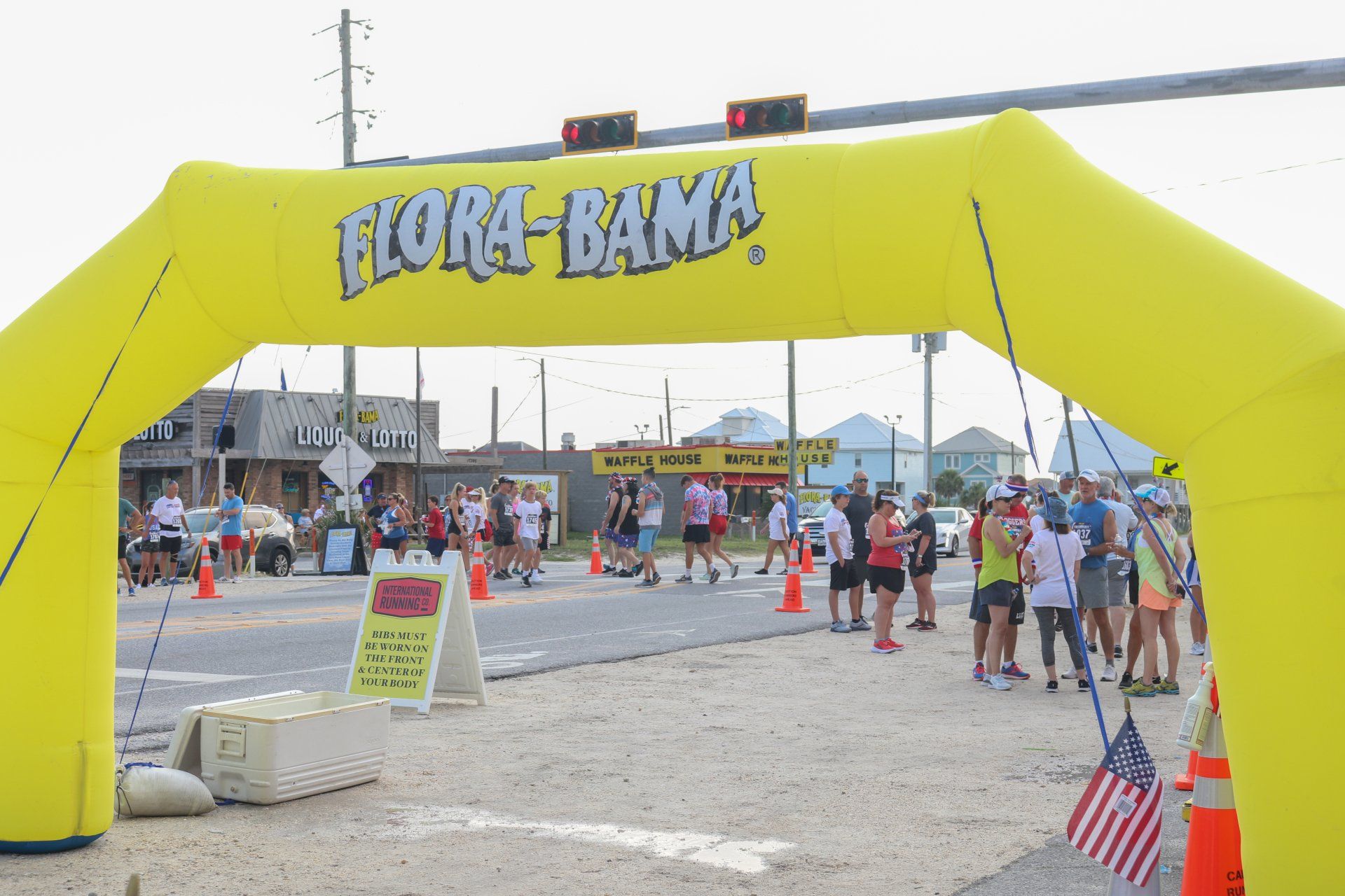 A yellow inflatable arch labeled Flora-Bama stands over a sandy area with people gathered near a coastal road and buildings.