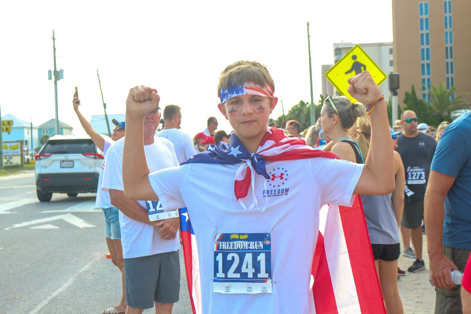 A runner wears a white T-shirt with bib 1241 and an American flag cape while flexing their arms outdoors.