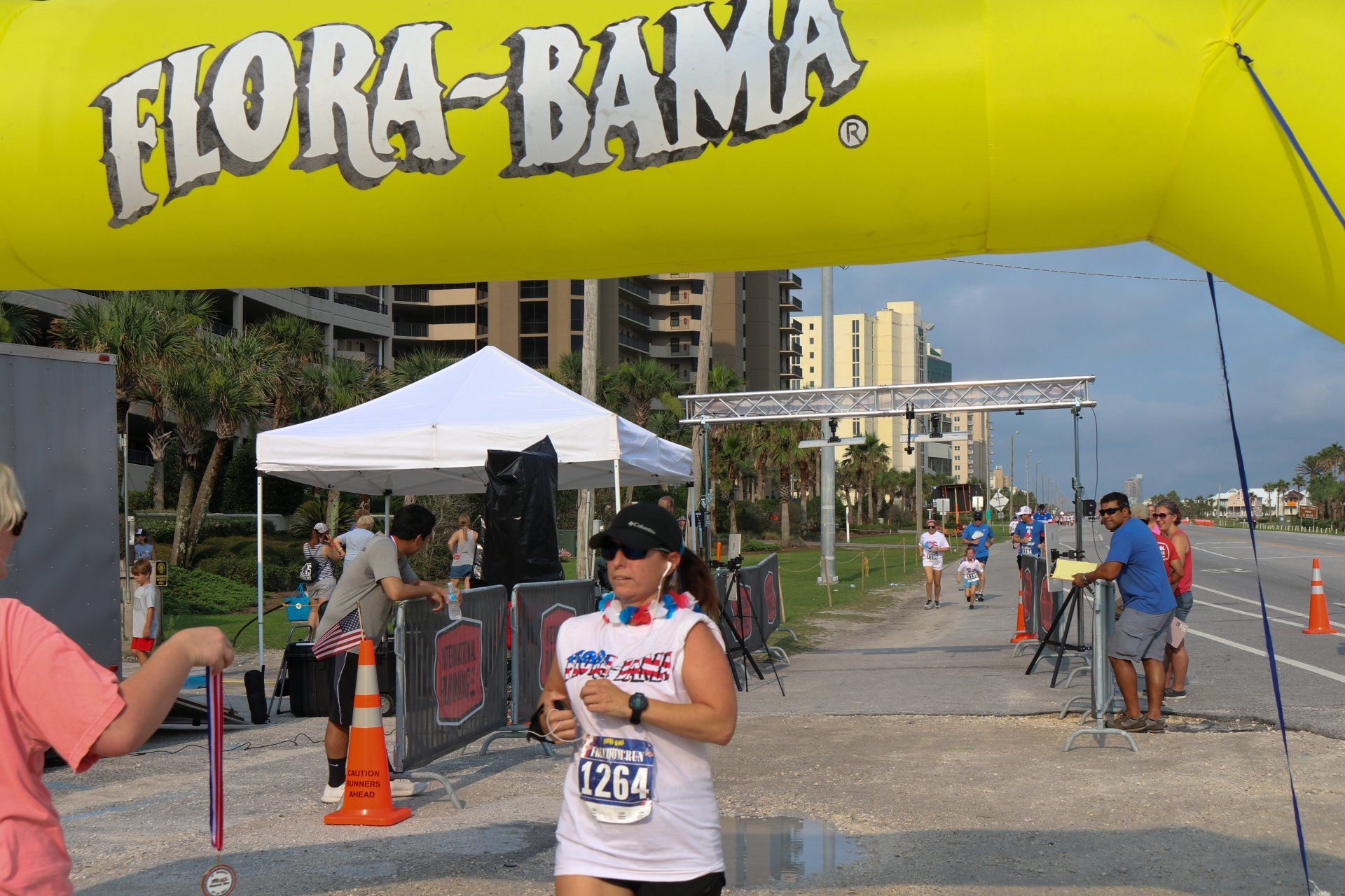 A runner with race bib 1264 crosses under a yellow Flora-Bama arch at an outdoor race event.