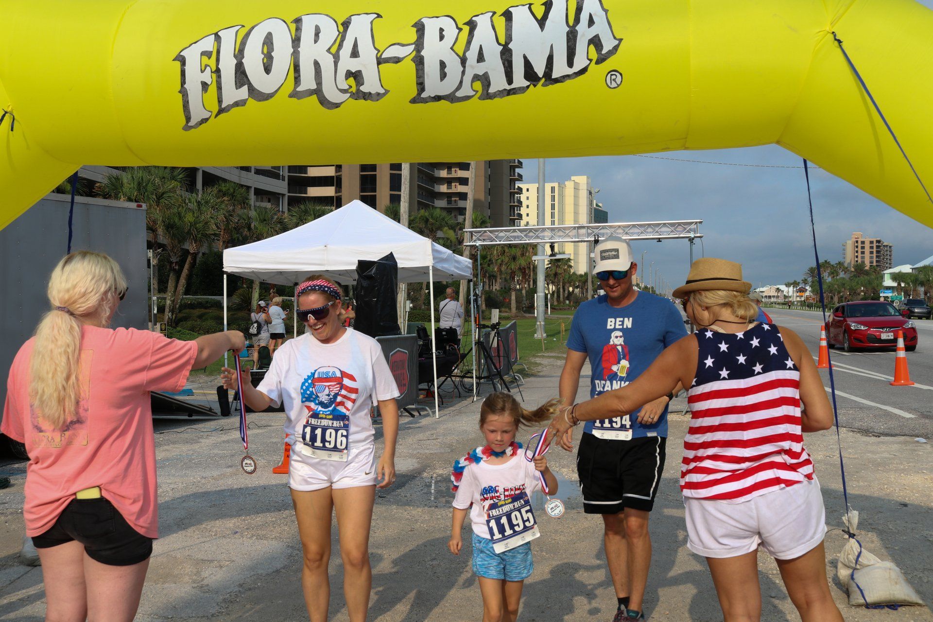 Participants cross under a yellow Flora-Bama archway at a race finish line with volunteers awarding medals.