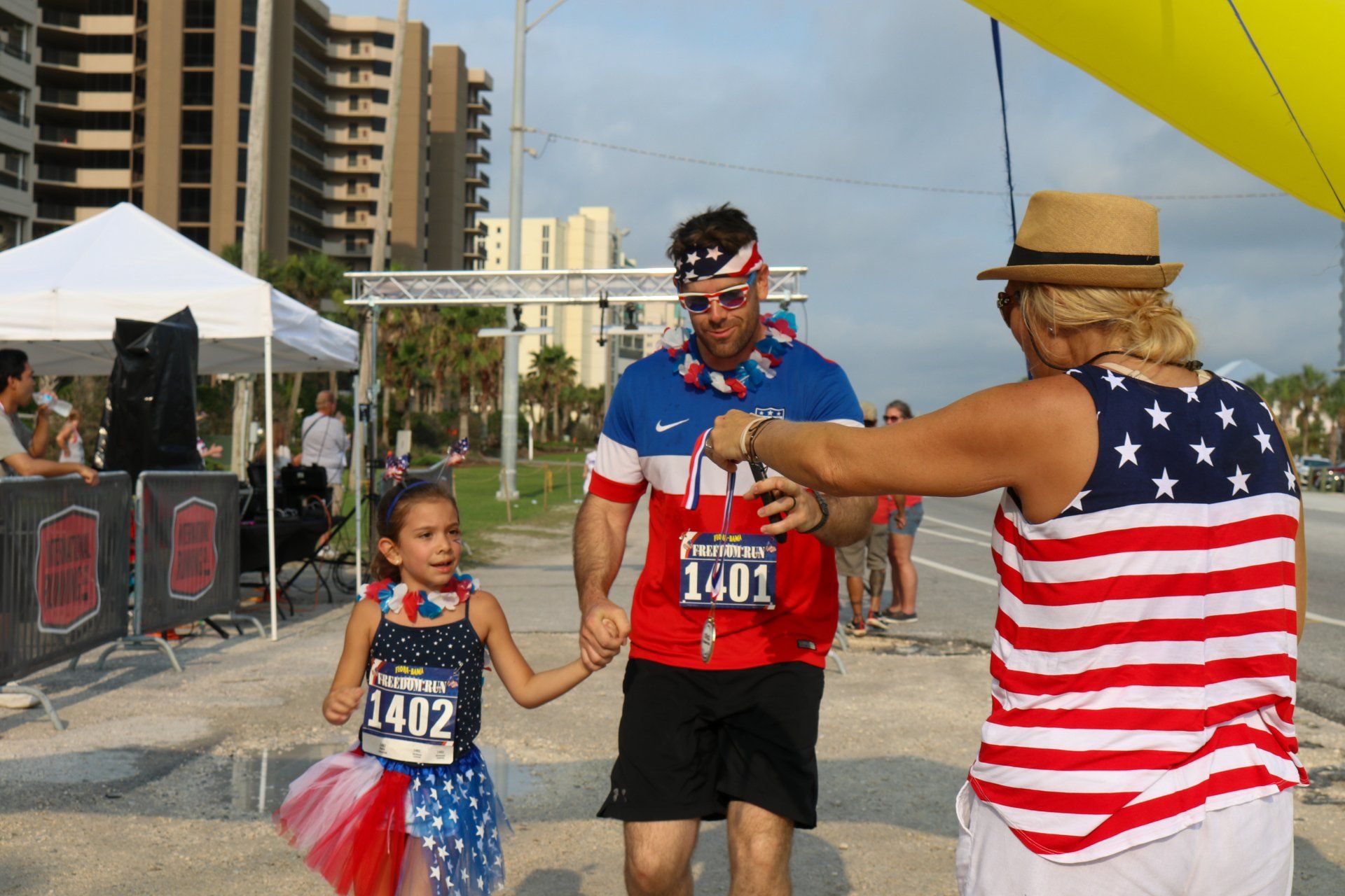 A race volunteer presents a finisher medal to a participant and child at an outdoor race with American-themed clothing.