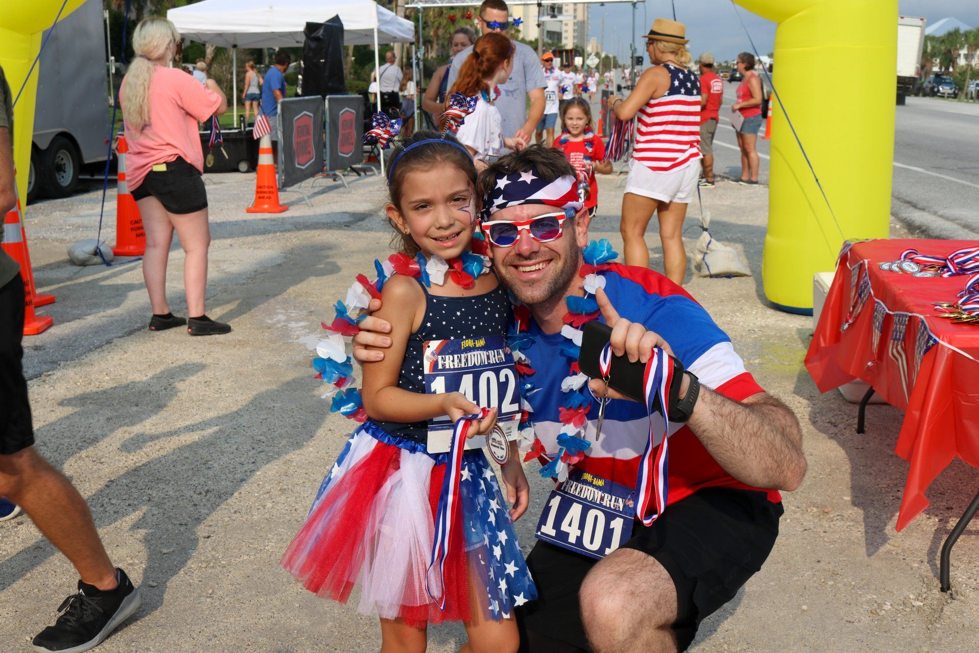 A man and a child wearing red, white, and blue race gear and leis pose for a photo at an outdoor event finish line.