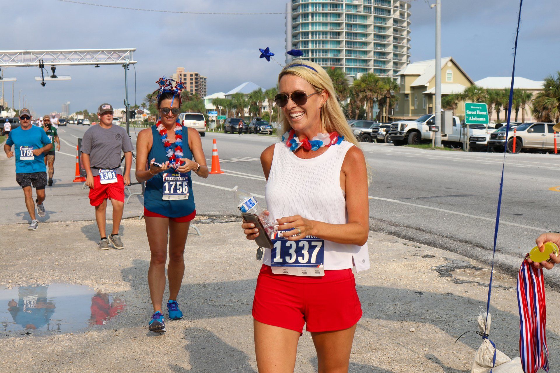Participants in a festive road race walk toward the camera, wearing red, white, and blue accessories and race bibs.