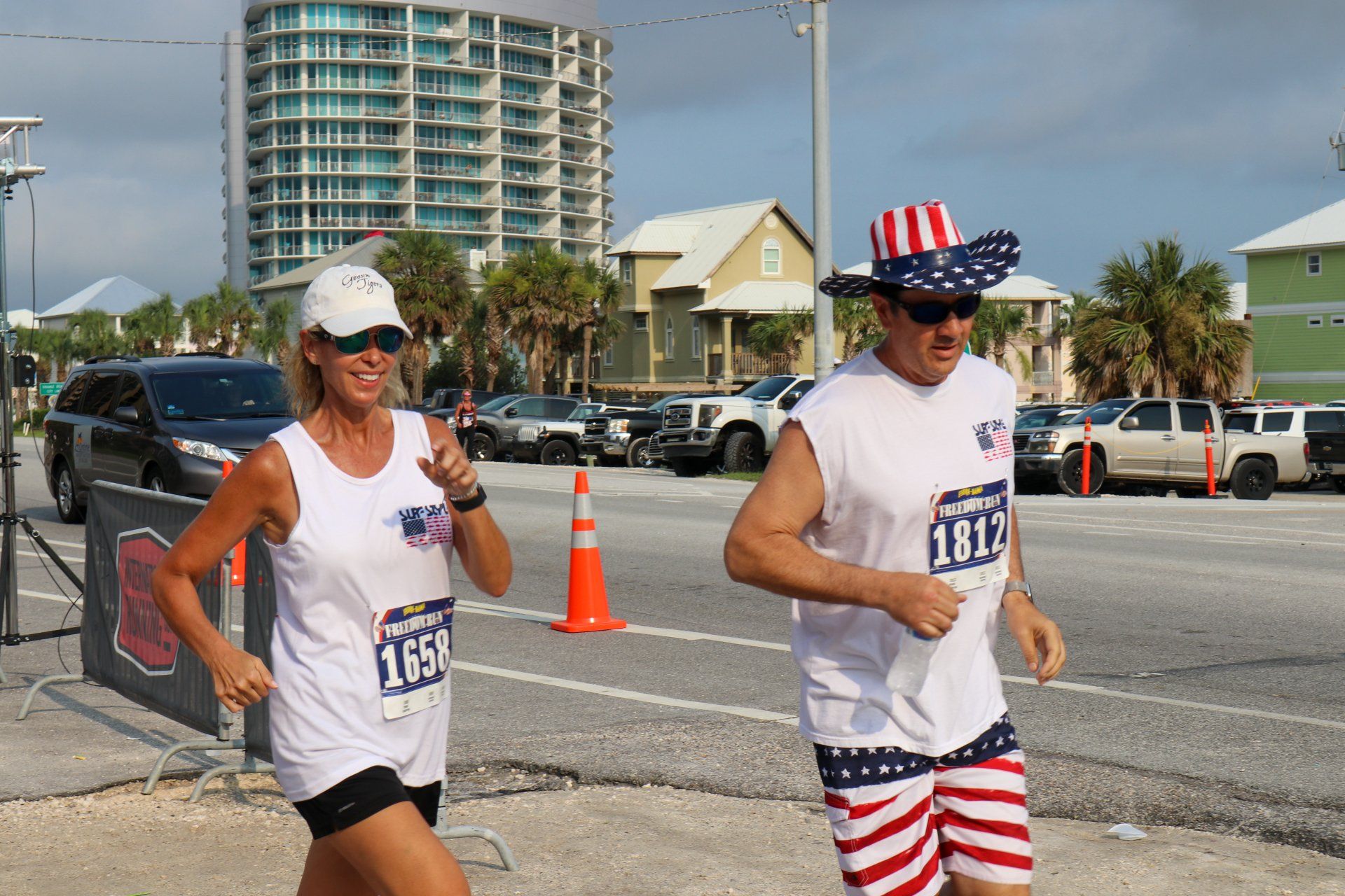 Two runners participate in a race wearing matching white shirts and race bibs, one in an American flag-themed hat and shorts.