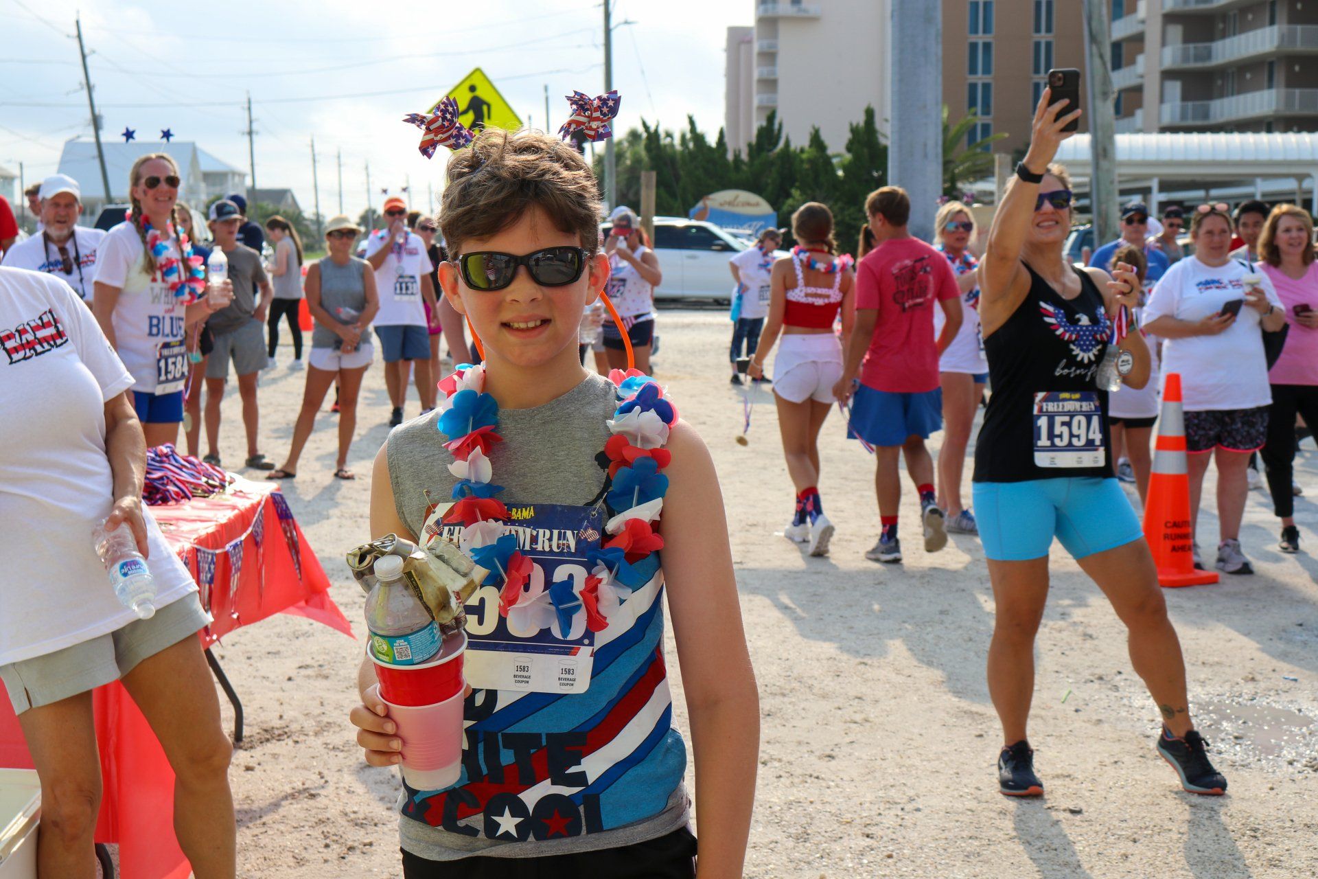 A person wearing sunglasses and patriotic accessories stands at a race, with runners and observers in the background.