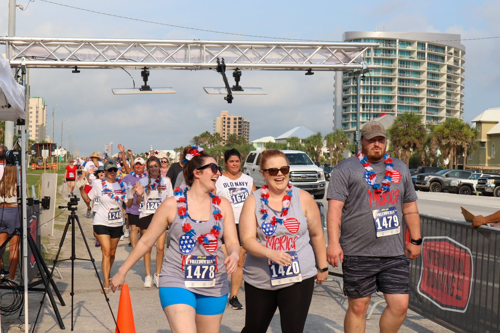Participants wearing festive leis and race bibs walk past a finish line archway at an outdoor event.