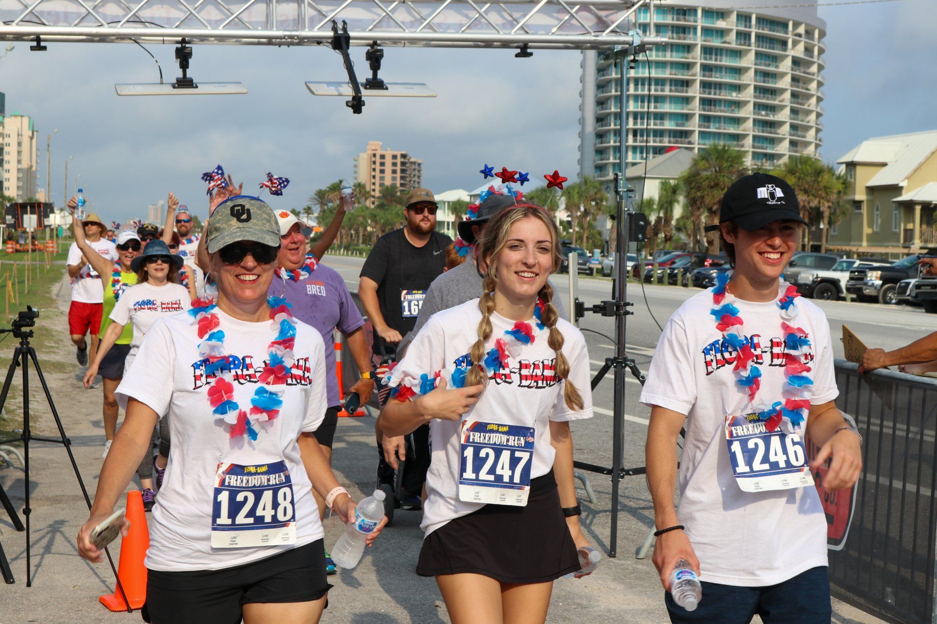 Three people wearing white race shirts with red, white, and blue leis smile while running outdoors at a race event.