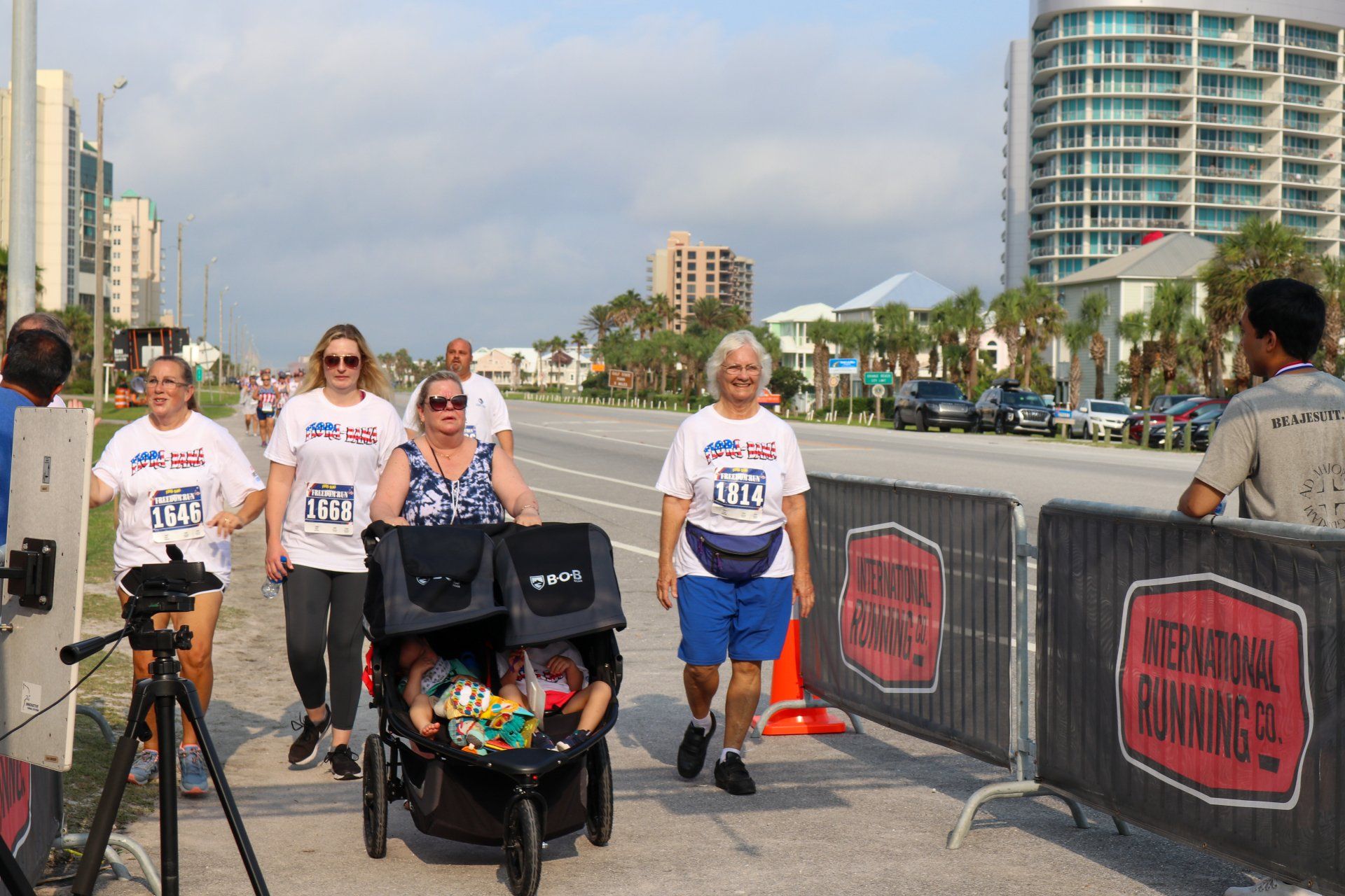 Four people in matching shirts walk on a path next to a road, one pushing a stroller with two small children.