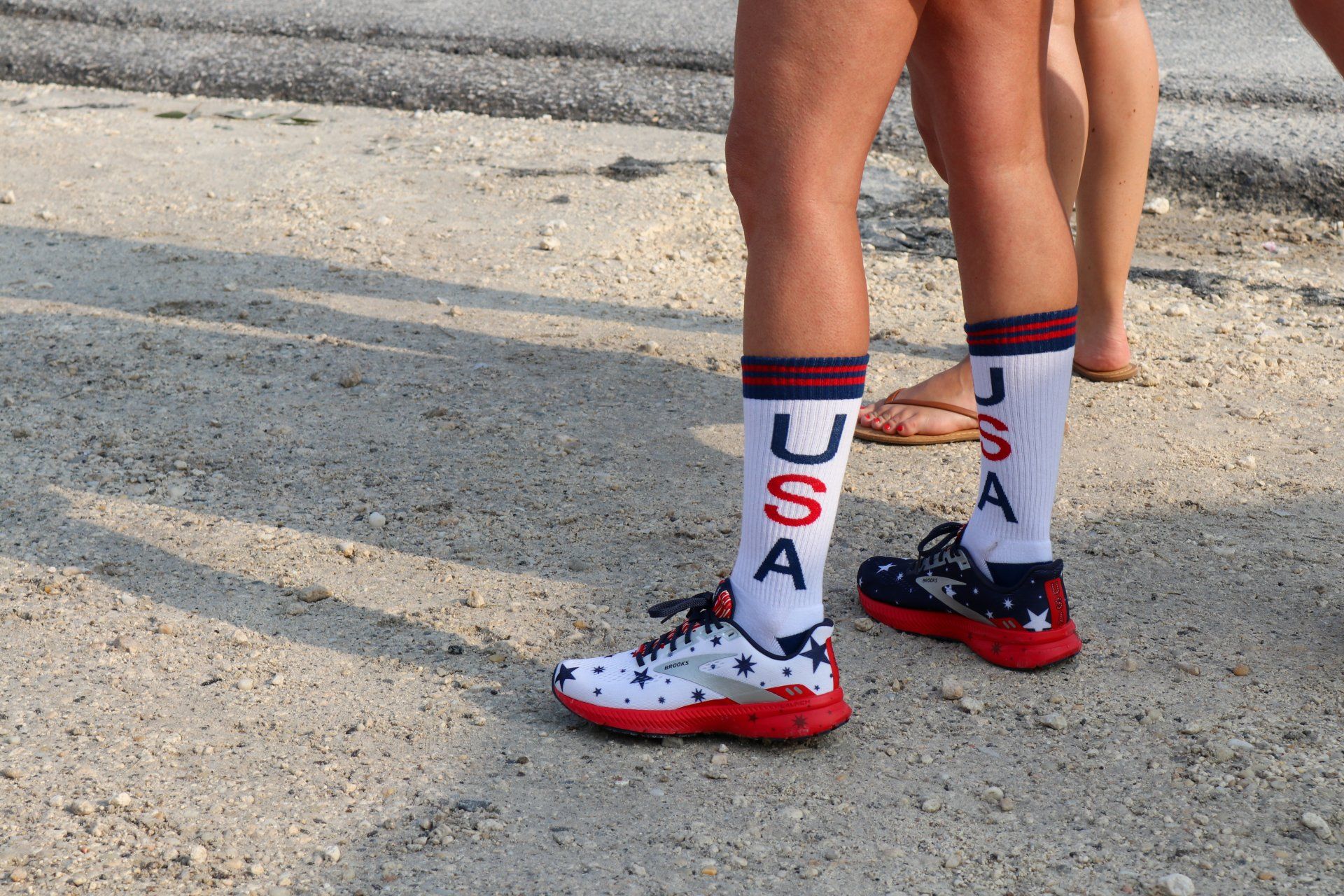 Person wearing white USA-themed socks and American flag-patterned athletic shoes standing on a gravel path.