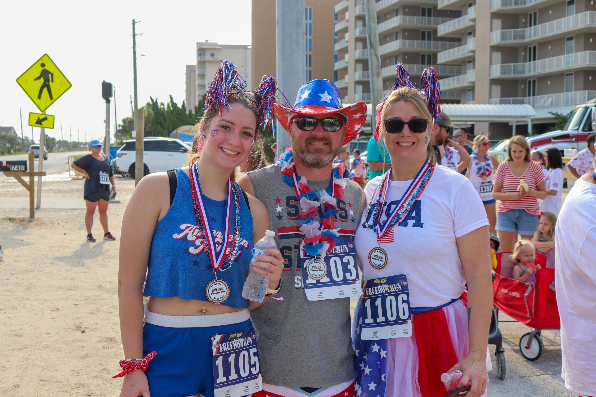 Three people wearing red, white, and blue race gear, medals, and festive hats pose for a photo at an outdoor race event.