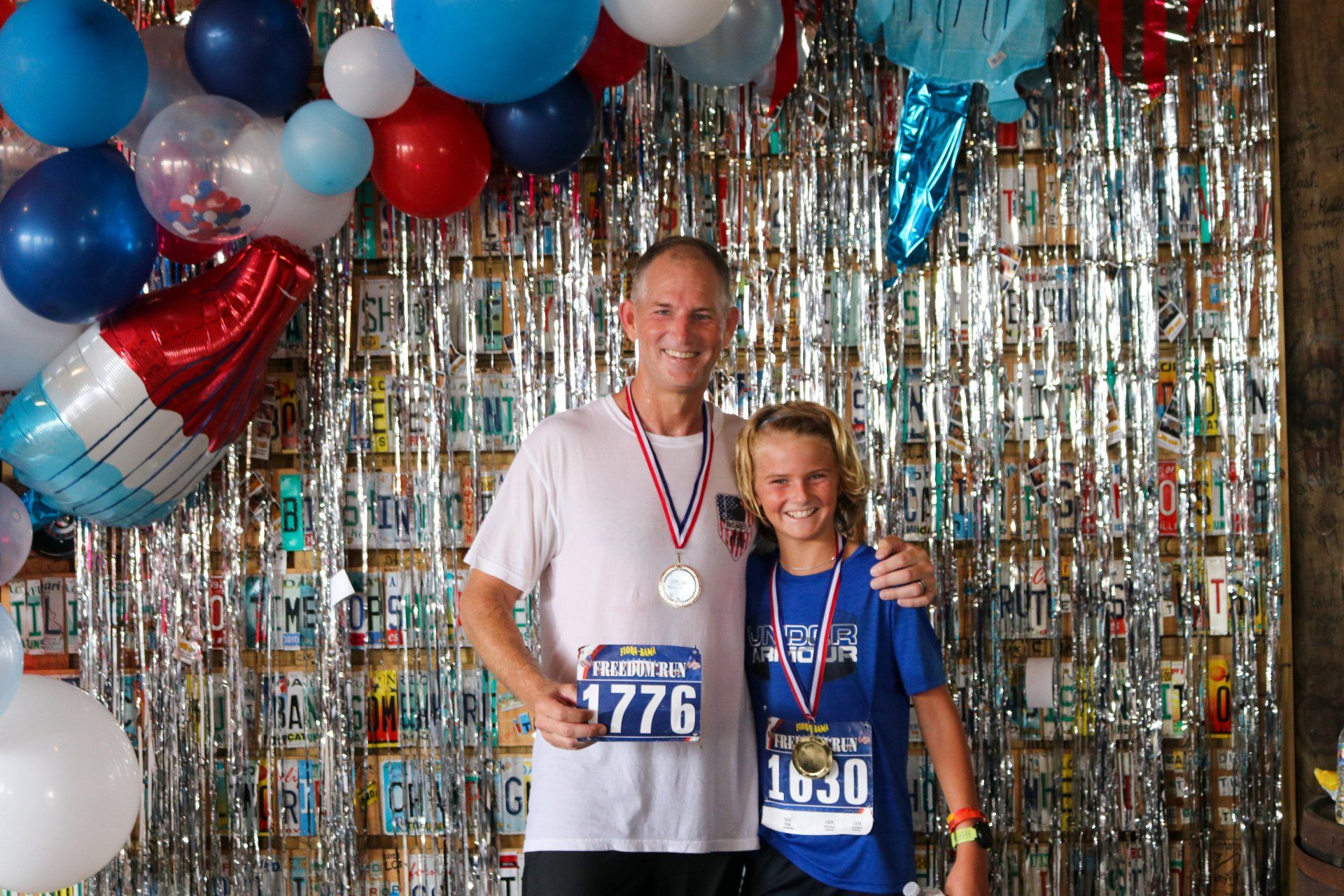 A smiling adult and youth wear medals and race bibs while posing together before a backdrop of balloons and silver fringe.