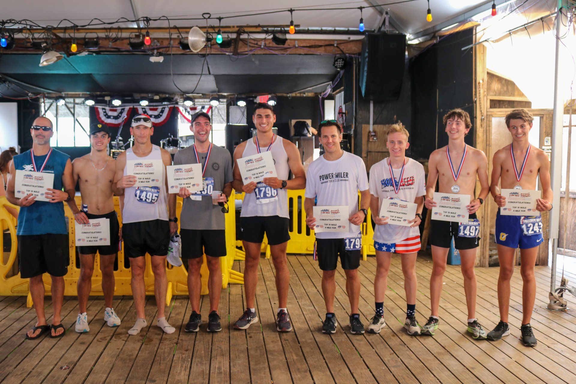 A line of nine people stand on a wooden deck holding race awards, wearing medals and athletic clothing.