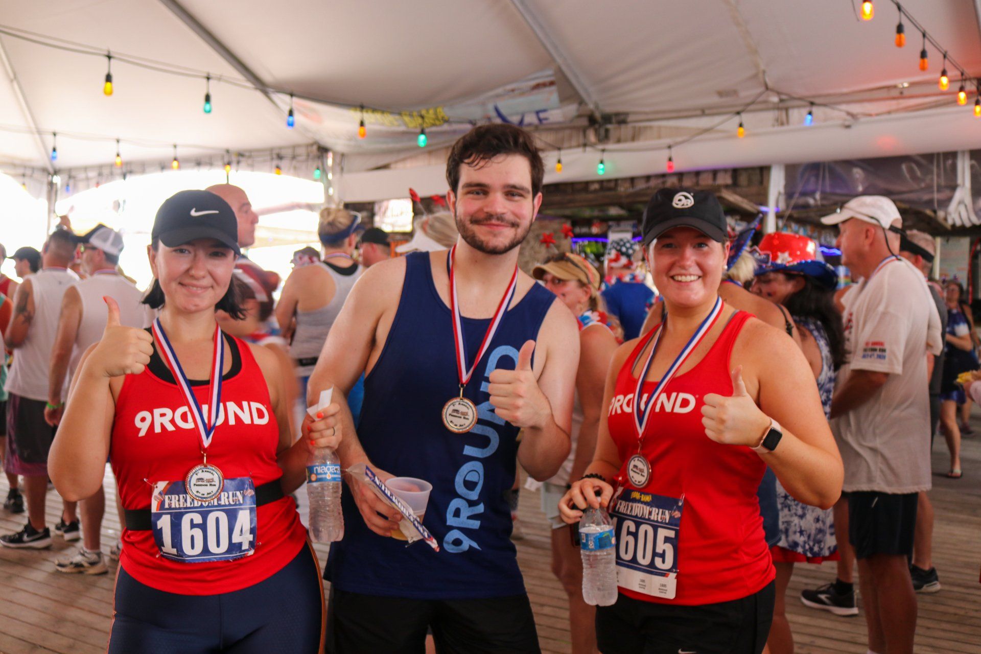 Three people wearing medals and 9Round gear smile and give thumbs up under a tent with decorative lights.