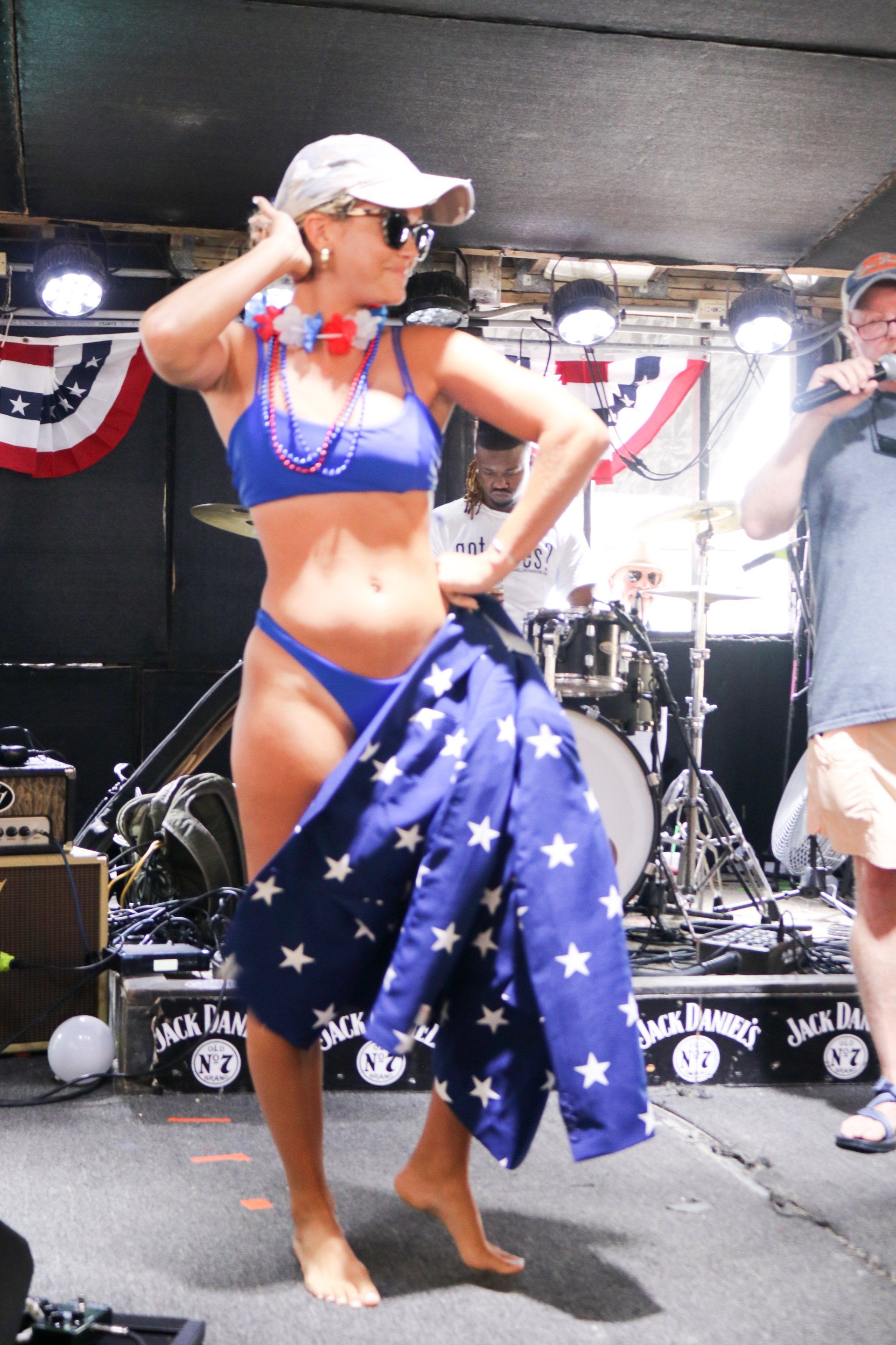 A person in a blue bikini and cap holds a star-patterned flag while standing on a stage at a Jack Daniel's event.