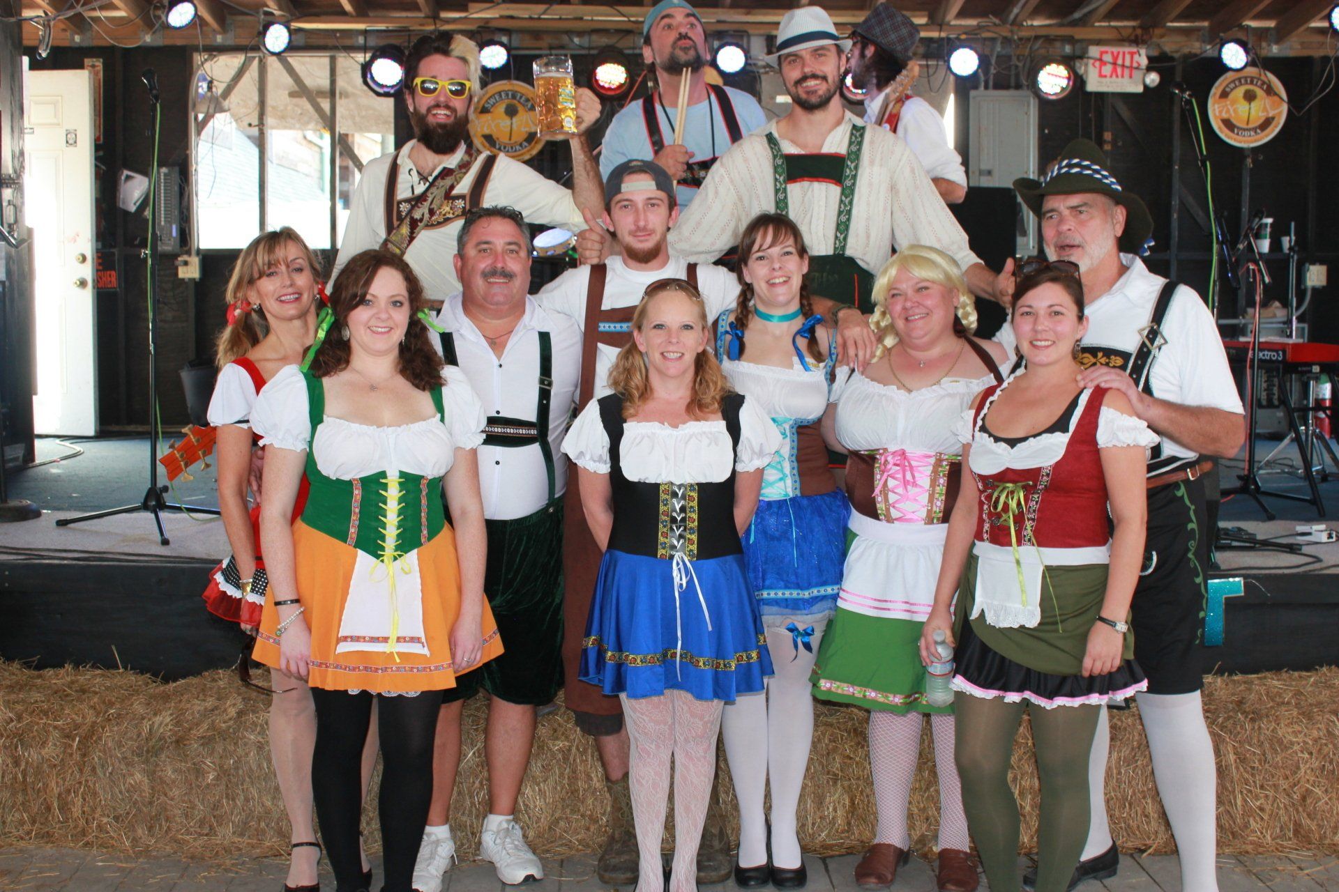 A group of people wearing traditional Bavarian Oktoberfest attire standing together indoors on a straw-covered stage.