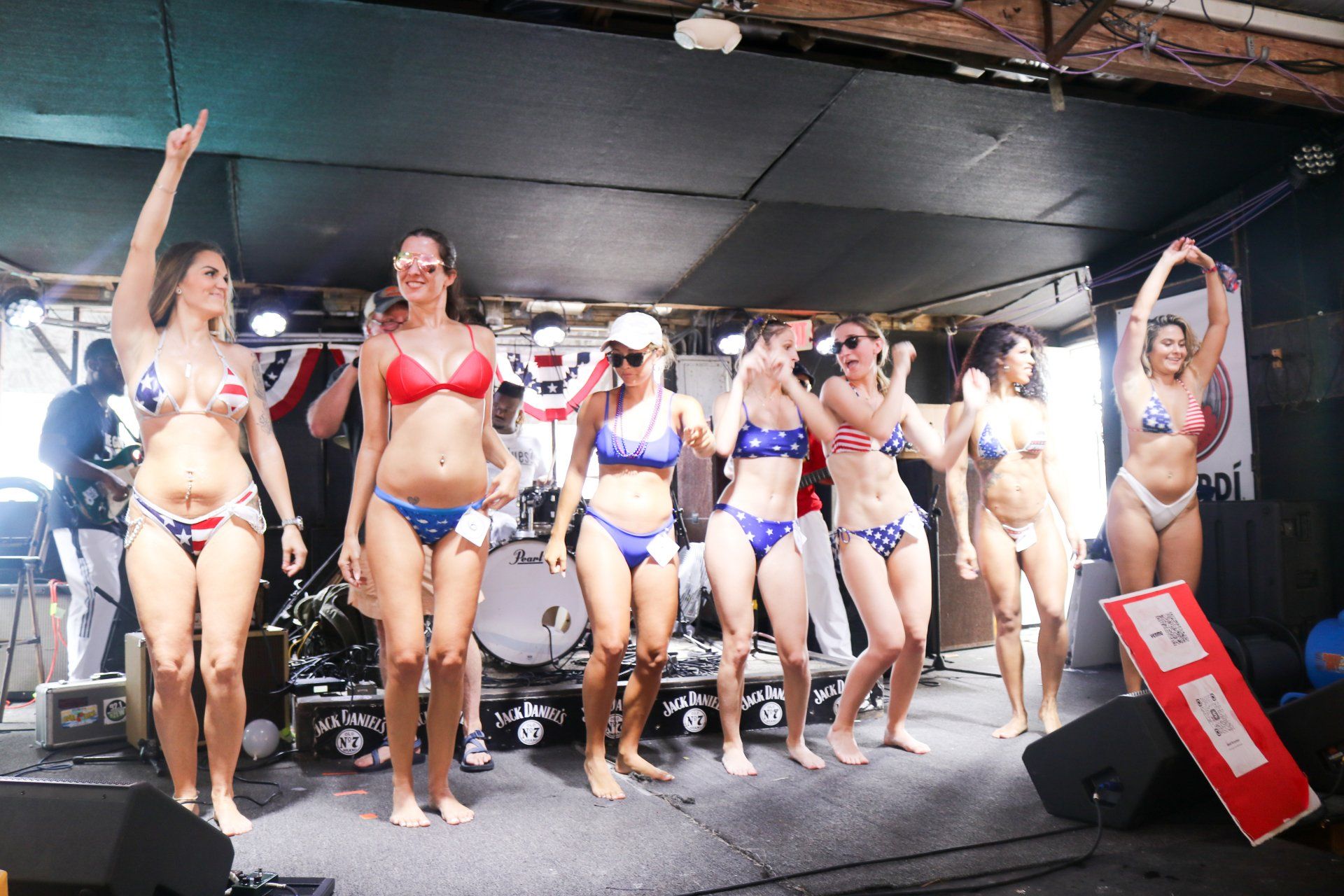Seven people in swimwear dance on a stage decorated with American flags, with a band and drum set visible behind them.
