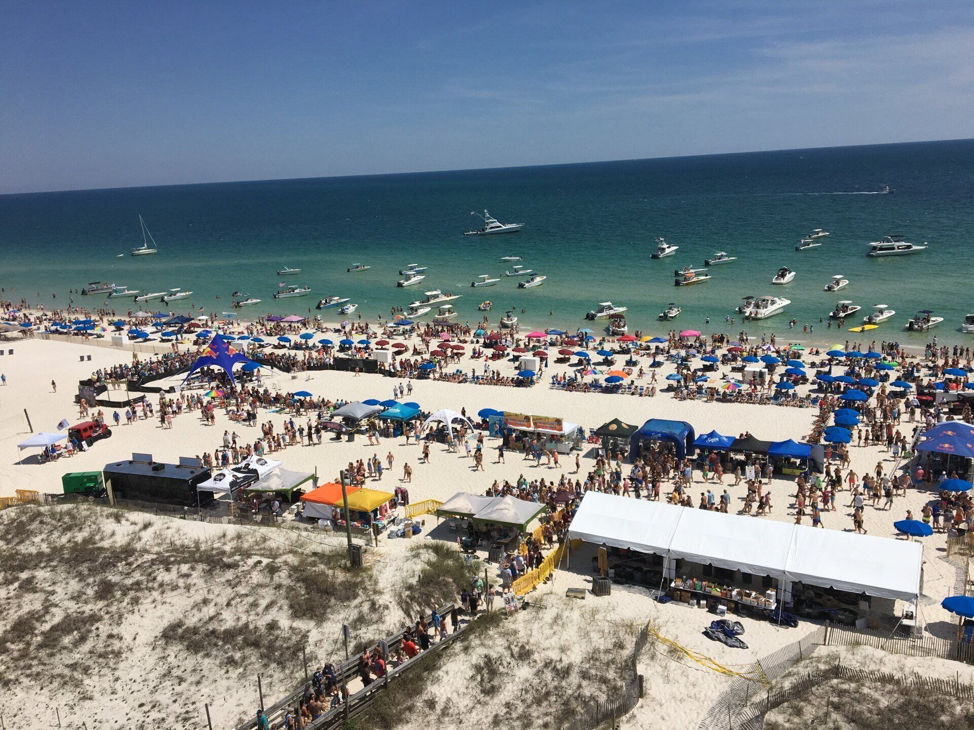 Aerial view of a crowded white sand beach with numerous boats anchored in the turquoise water and tents on the shore.