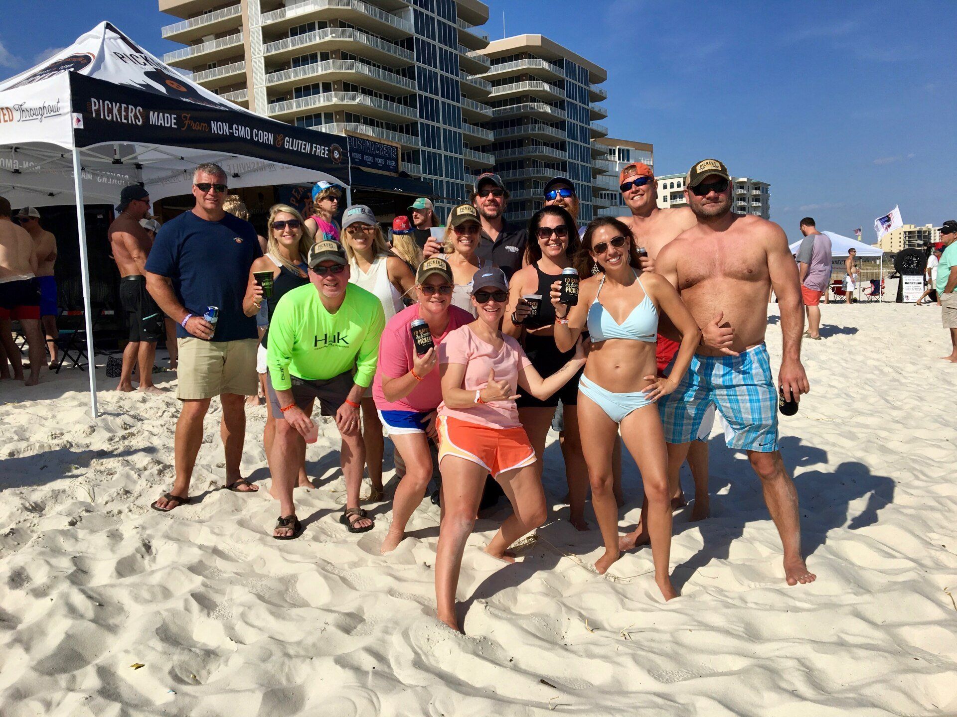 A large group of people stand together on a sandy beach in front of a white tent, smiling for a photo.