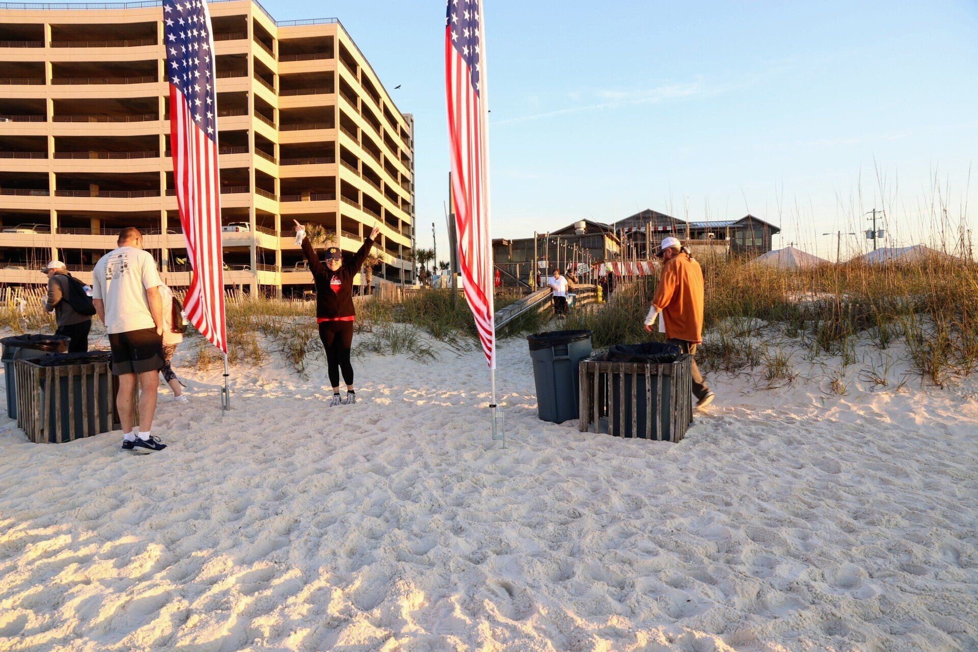 People clean a sandy beach near trash bins and two American flags, with a large building under construction in back.