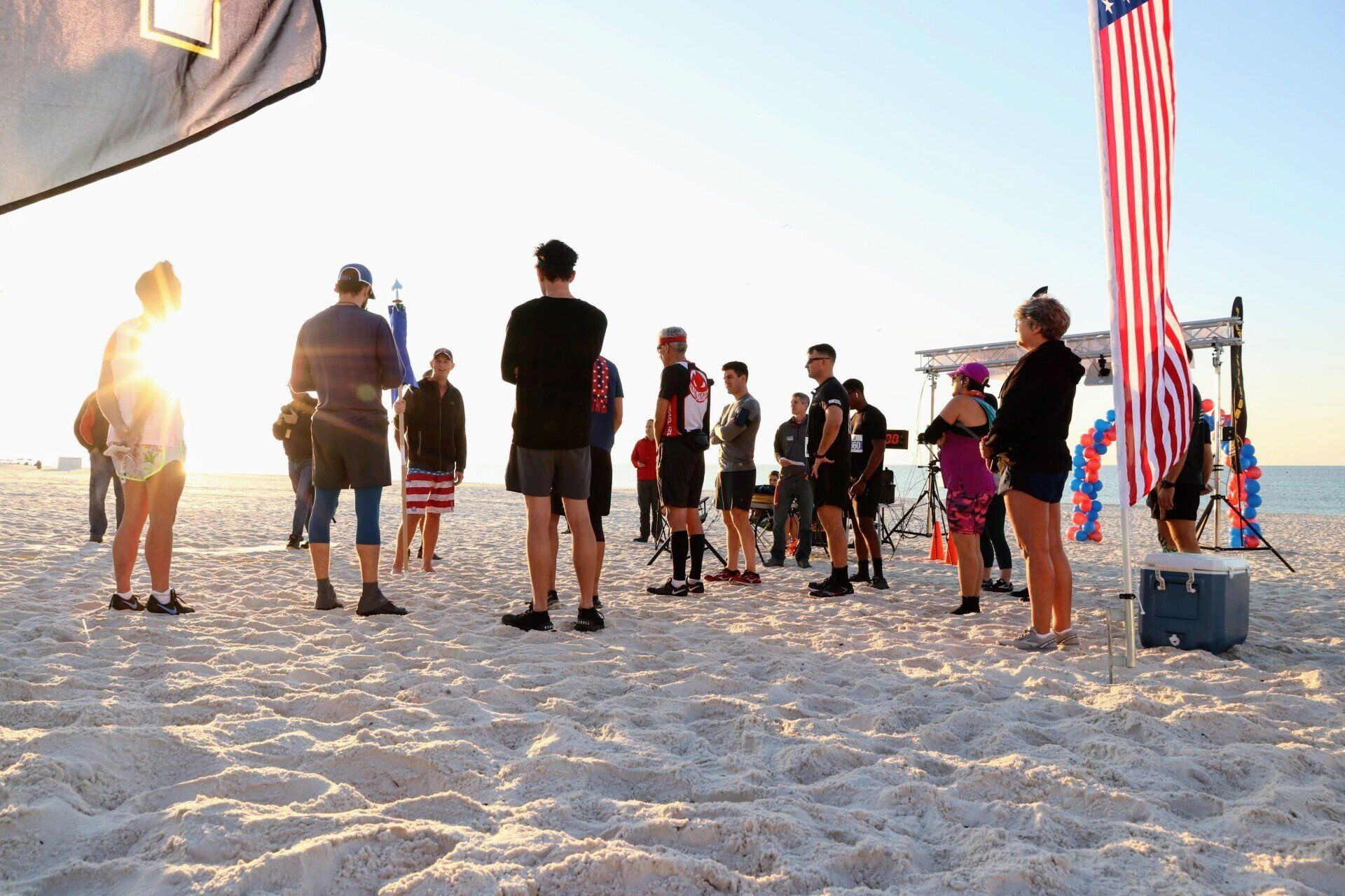 A group of people standing on a sandy beach at sunset, with a large American flag standing on a pole to the right.
