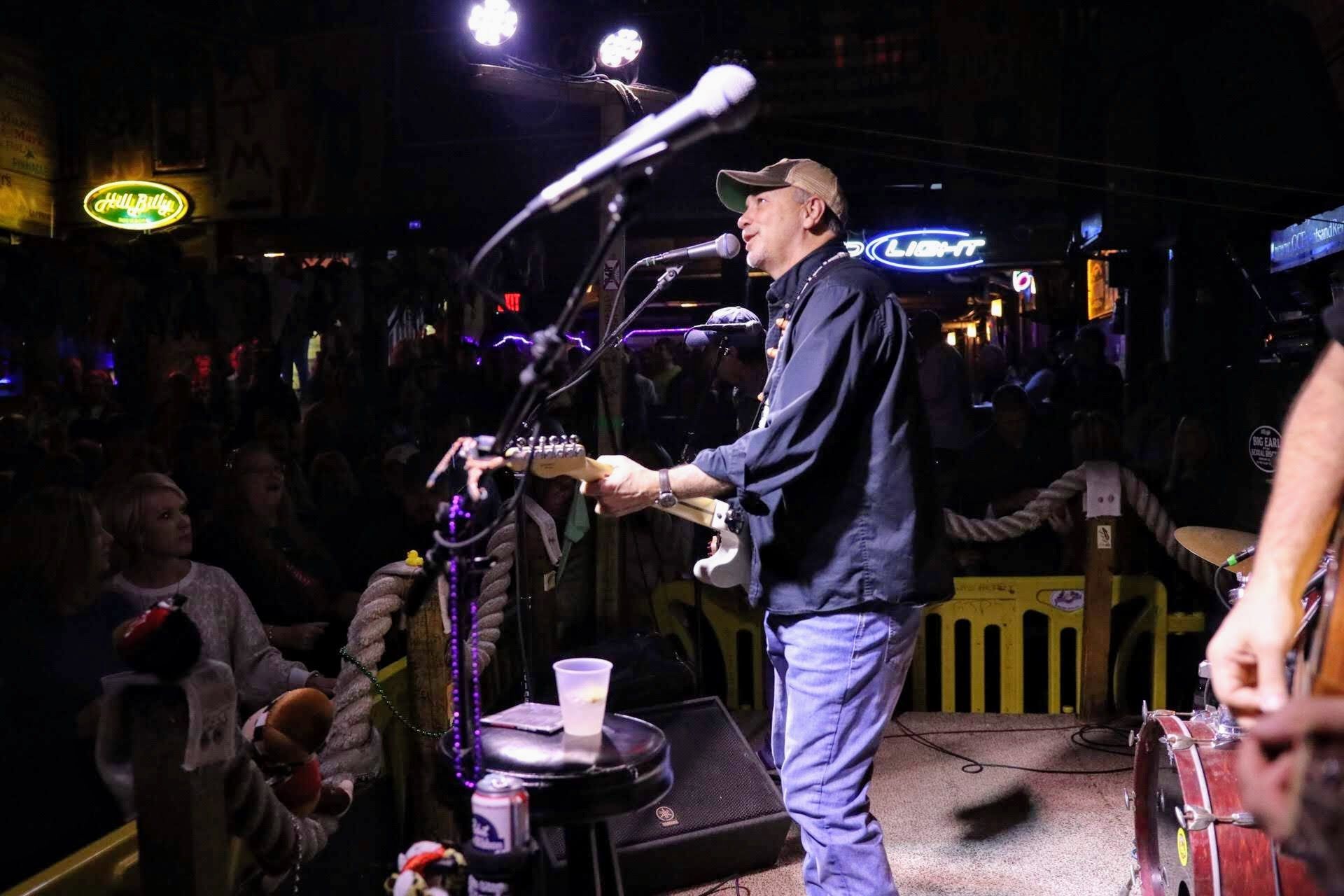 A musician wearing a cap performs on stage, singing into a microphone while playing an electric guitar at a bar.