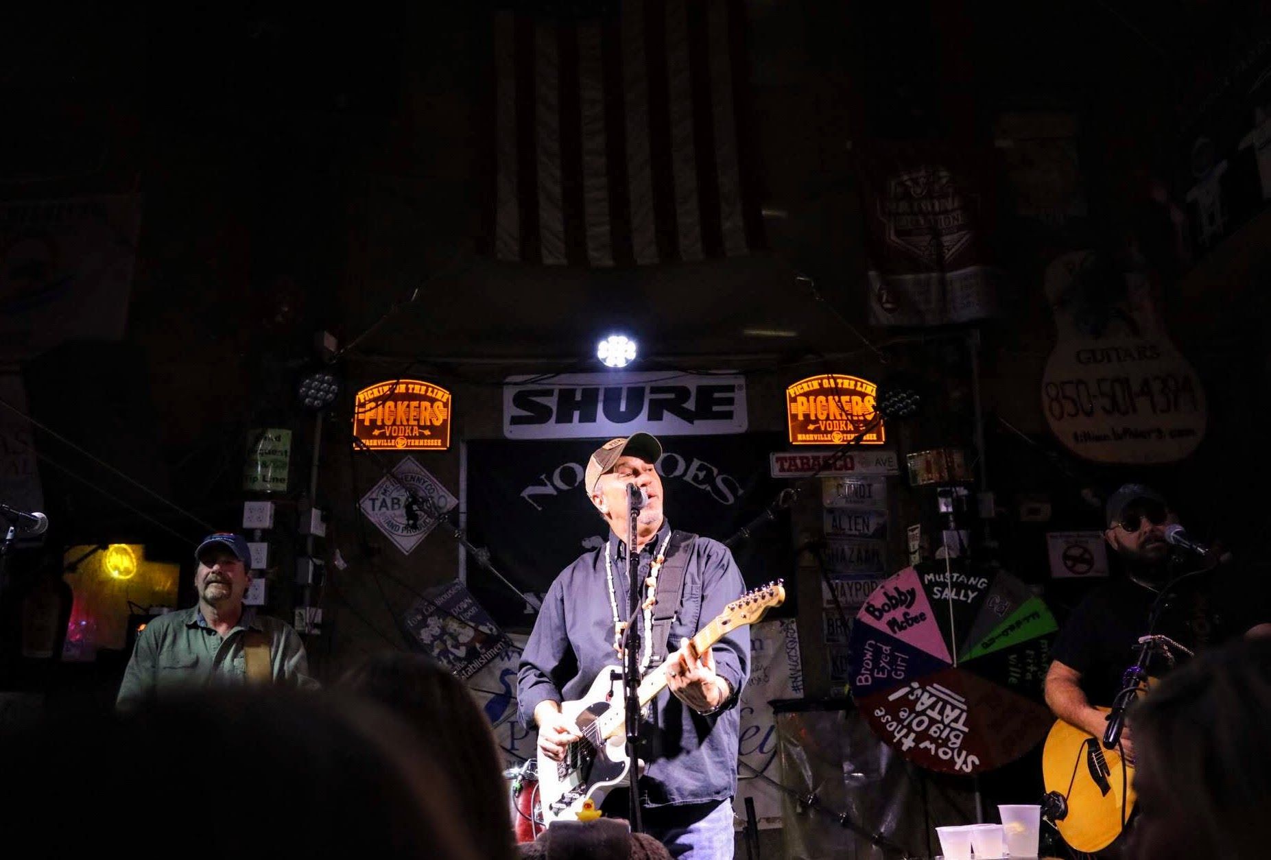 Three musicians perform live on a dimly lit stage decorated with various signs and an American flag.