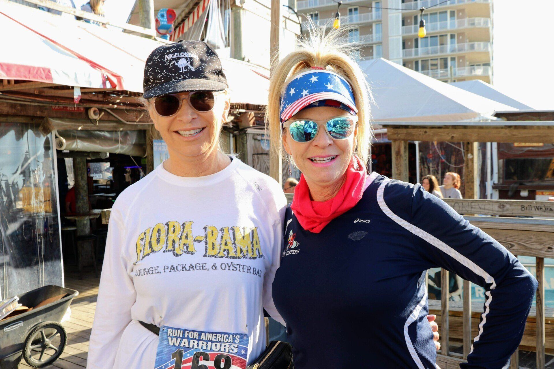 Two people in hats and sunglasses pose smiling in front of a wooden building, wearing race bibs.